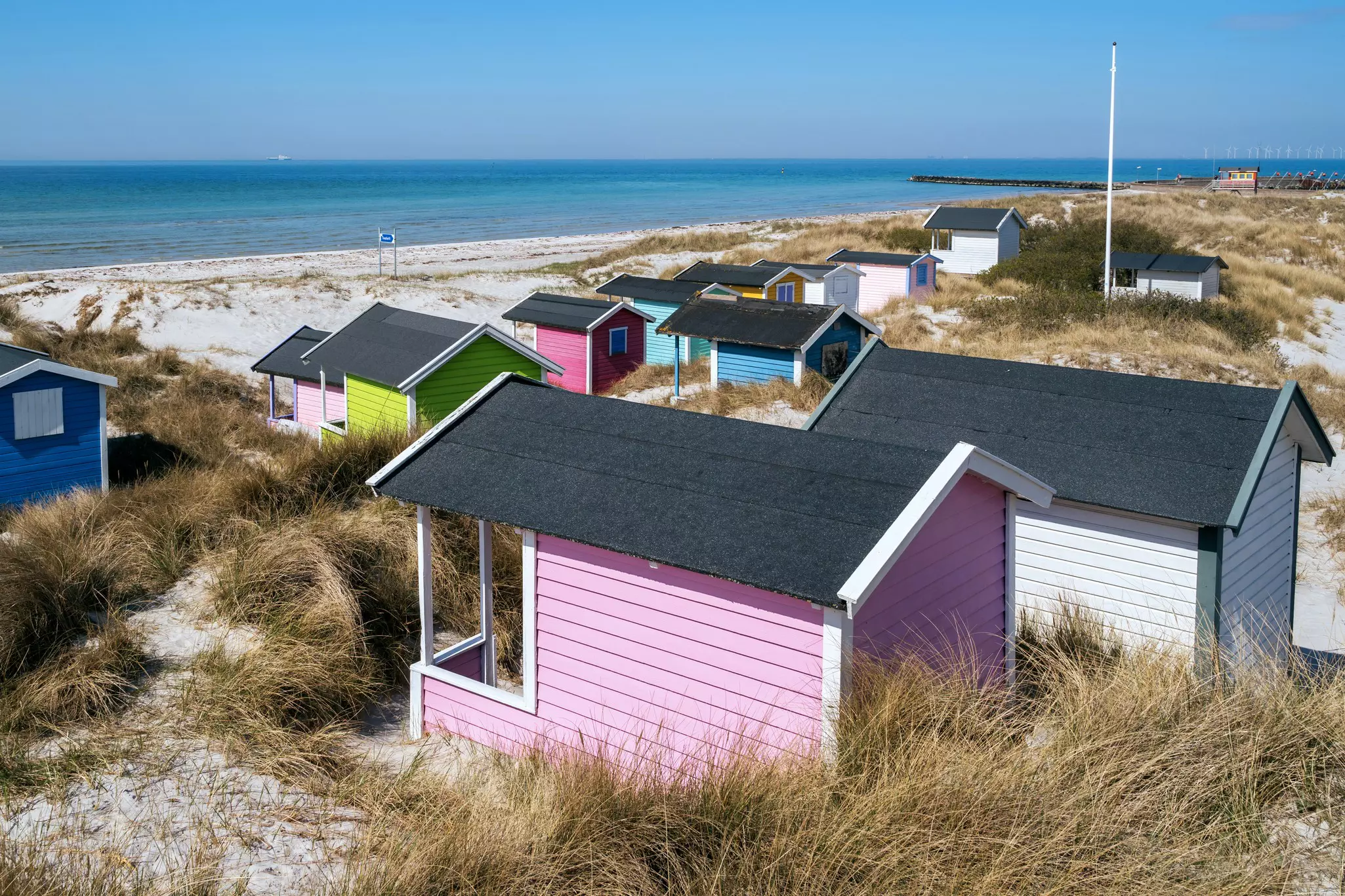 Pastel-colored wooden beach huts among sand dunes facing out towards the sea.