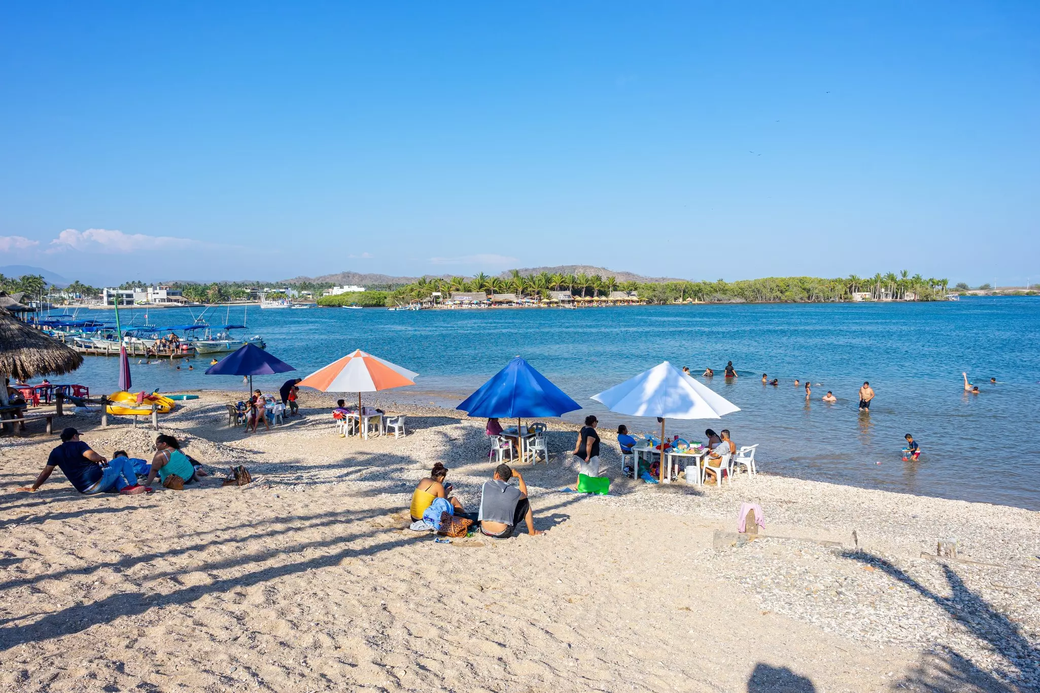 Families sit under umbrellas at a beach with a shallow swimming area