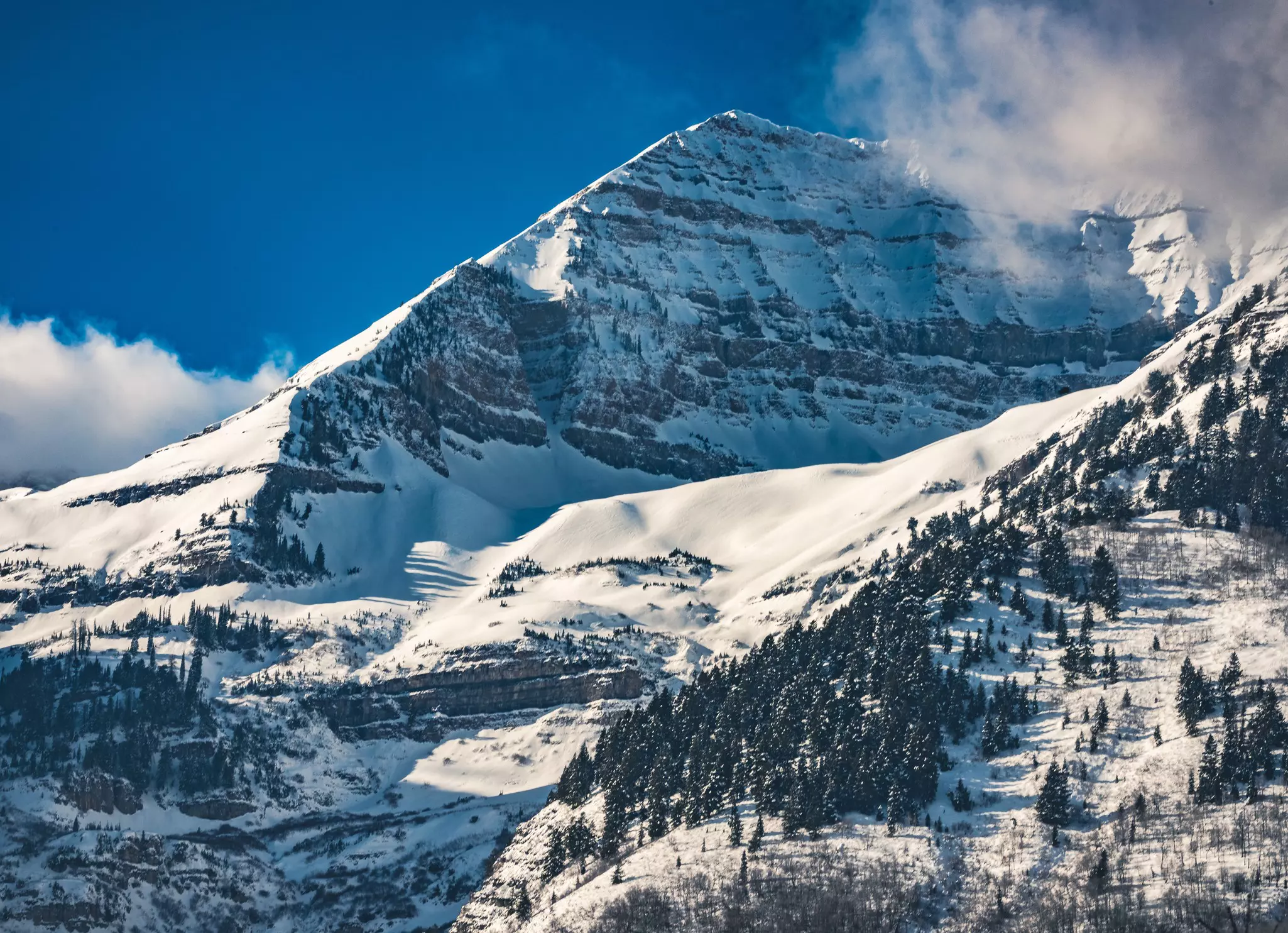 A peak with steep cliffs is covered with snow, as clouds grace the top.