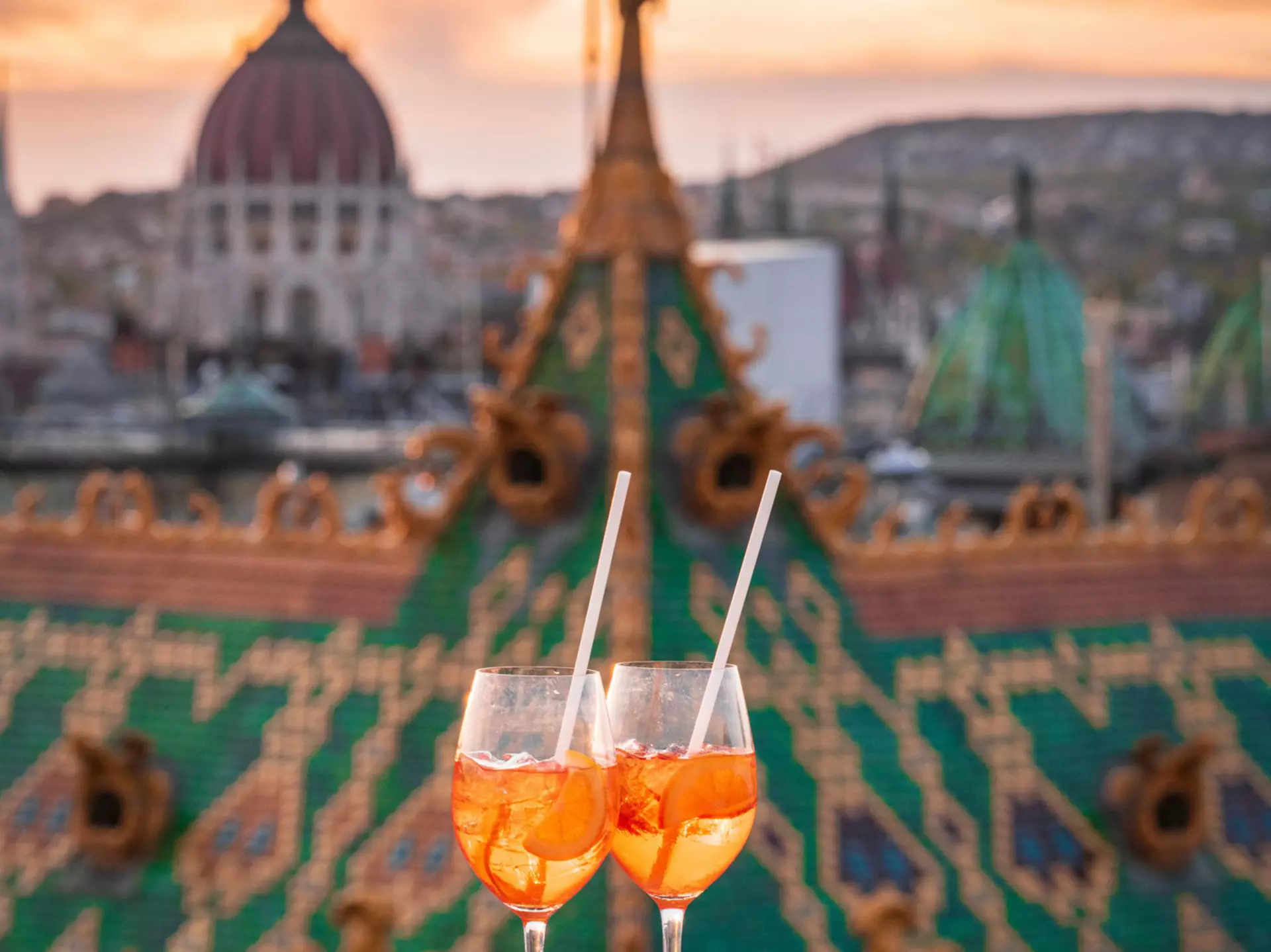A pair of cocktails at sunset on a rooftop ledge in focus, with buildings including the Parliament in the background, Budapest, Hungary, Europe