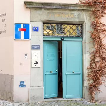 Pale pink old-town building with open turquoise double doors under an inscribed stone lintel and a blue bicycle