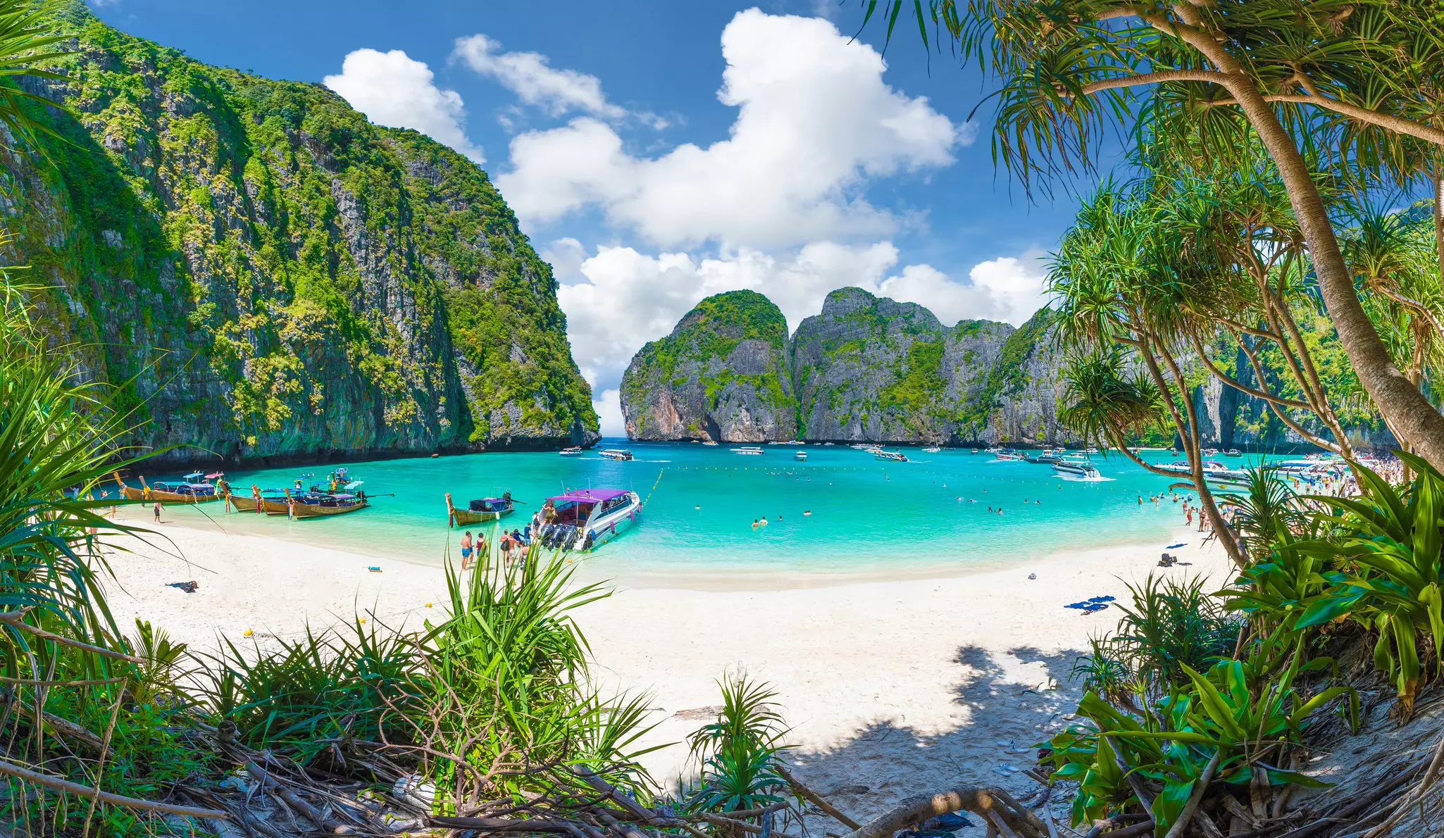 A white sand beach seen from a shaded area; boats are moored in the turquoise cove, and tree-covered rock formations enclose the cove