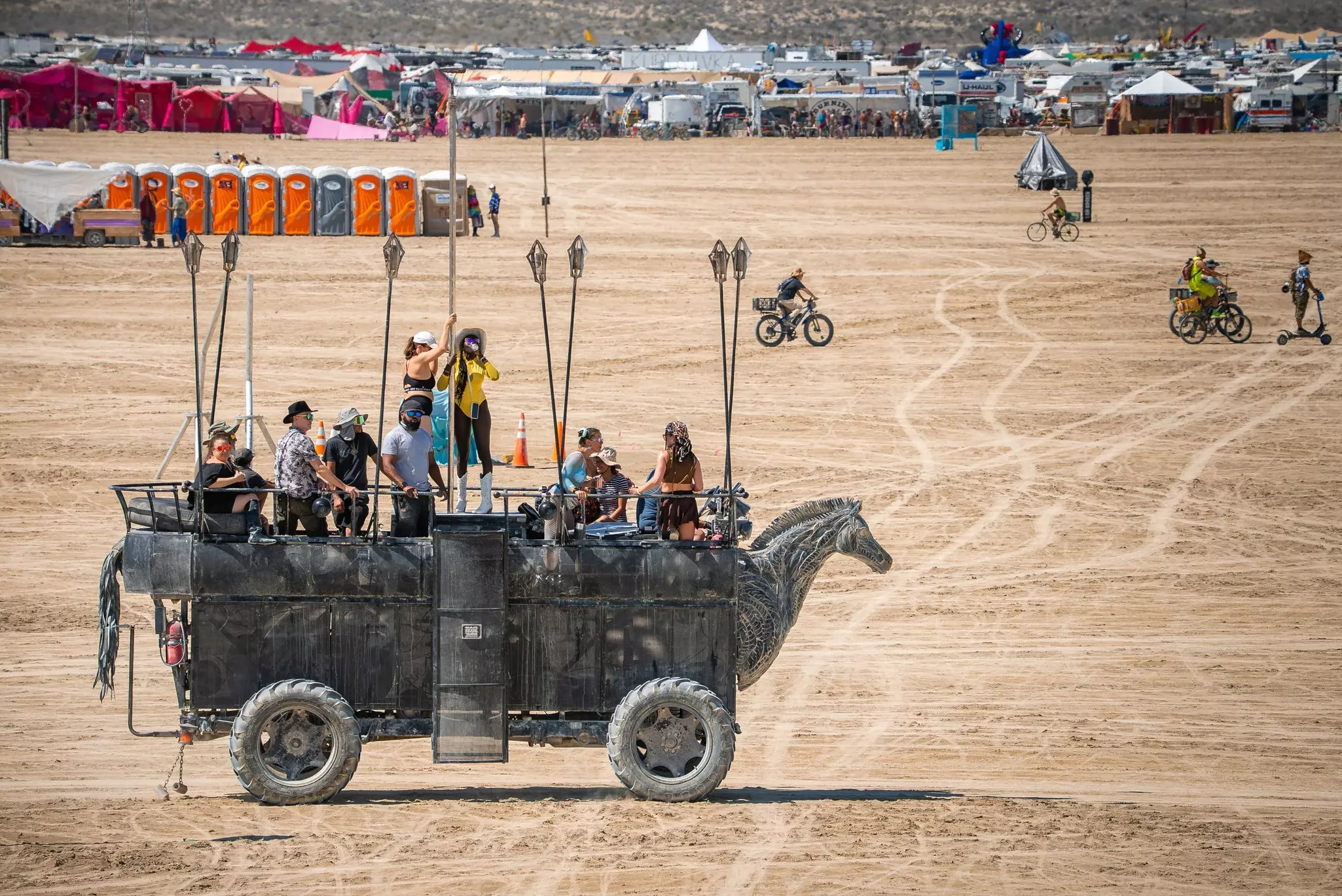 A carriage-style vehicle in the desert with tents clustered in the background. A line of portable toilets is off to the left of the photo