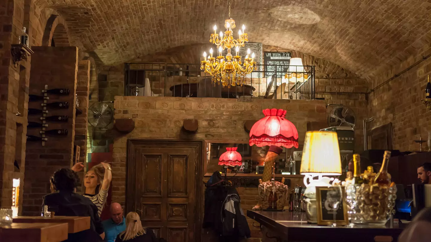 A cosy, cellar-like room with brick ceiling is lit by a chandelier and two standing lamps. Wine bottles can be seen on the bar to the right.