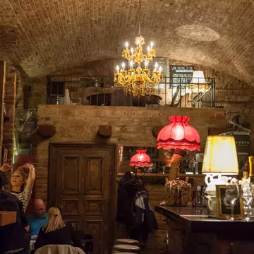A cosy, cellar-like room with brick ceiling is lit by a chandelier and two standing lamps. Wine bottles can be seen on the bar to the right.