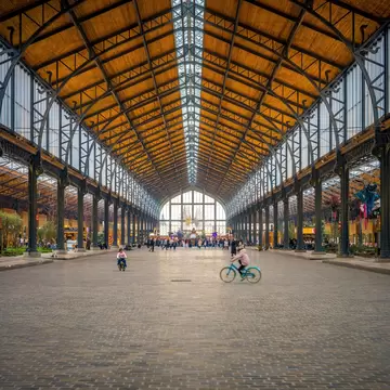 Long cobbled hall under a wooden vaulted roof and skylight, lined with iron columns and shops, a child cycling in front.