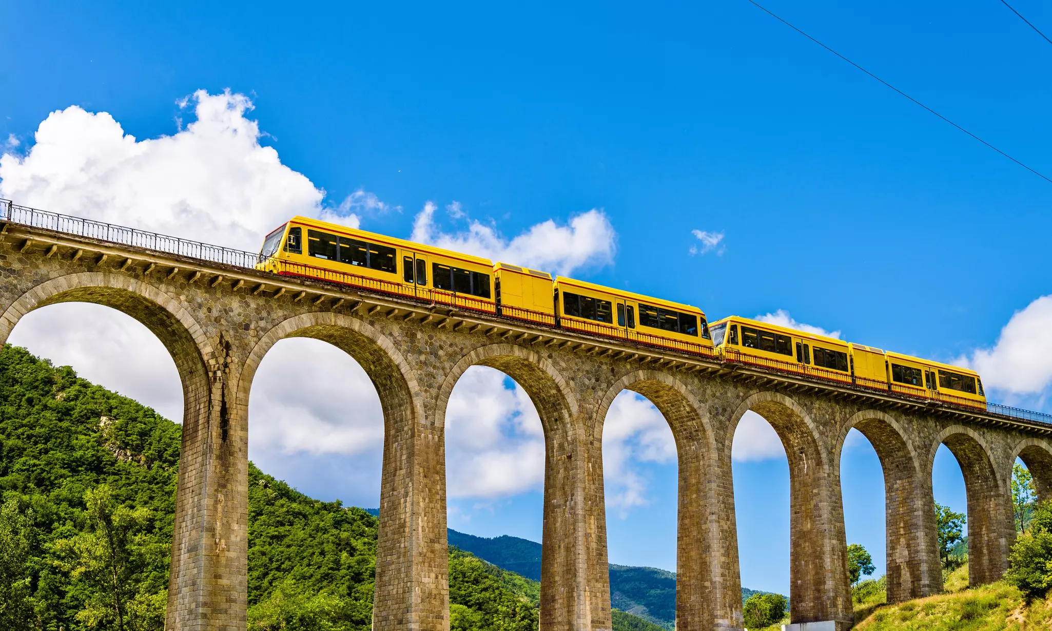A bright yellow train goes over a high bridge with blue sky and clouds in the background