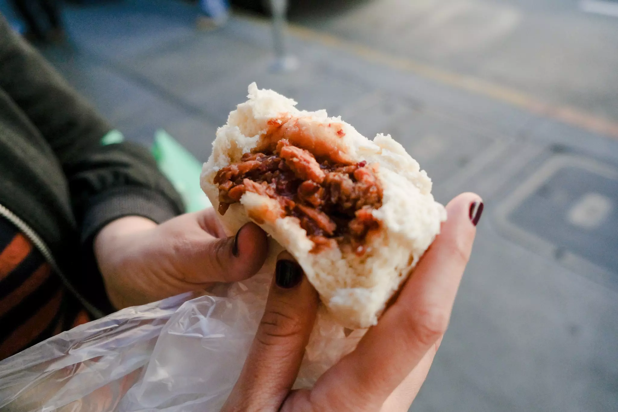 Nail-polished hands hold a pale bun filled with pork and sauce
