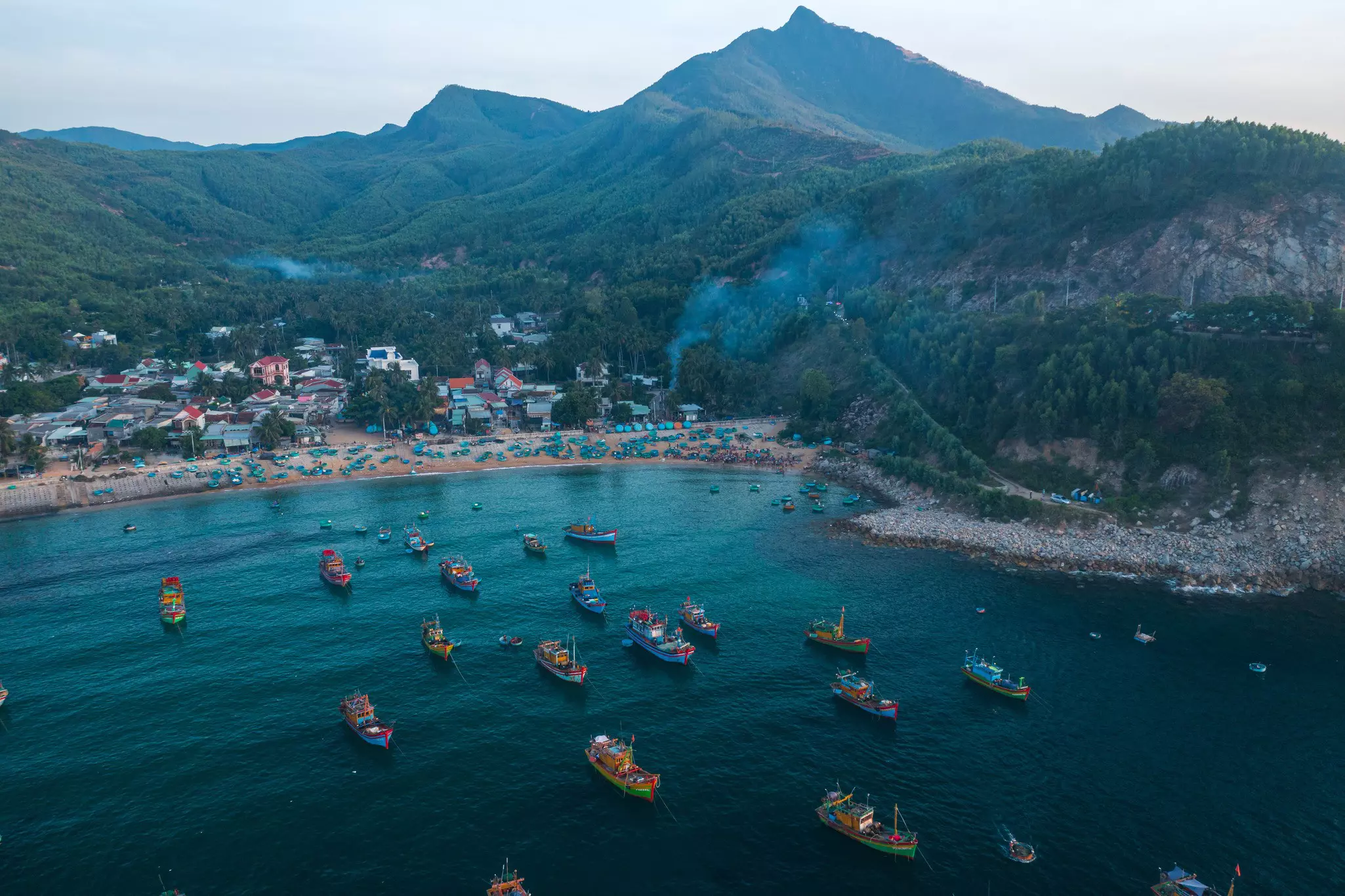 Colorful fishing boats moored in a cove. The beach is full of people in the early morning gloom.