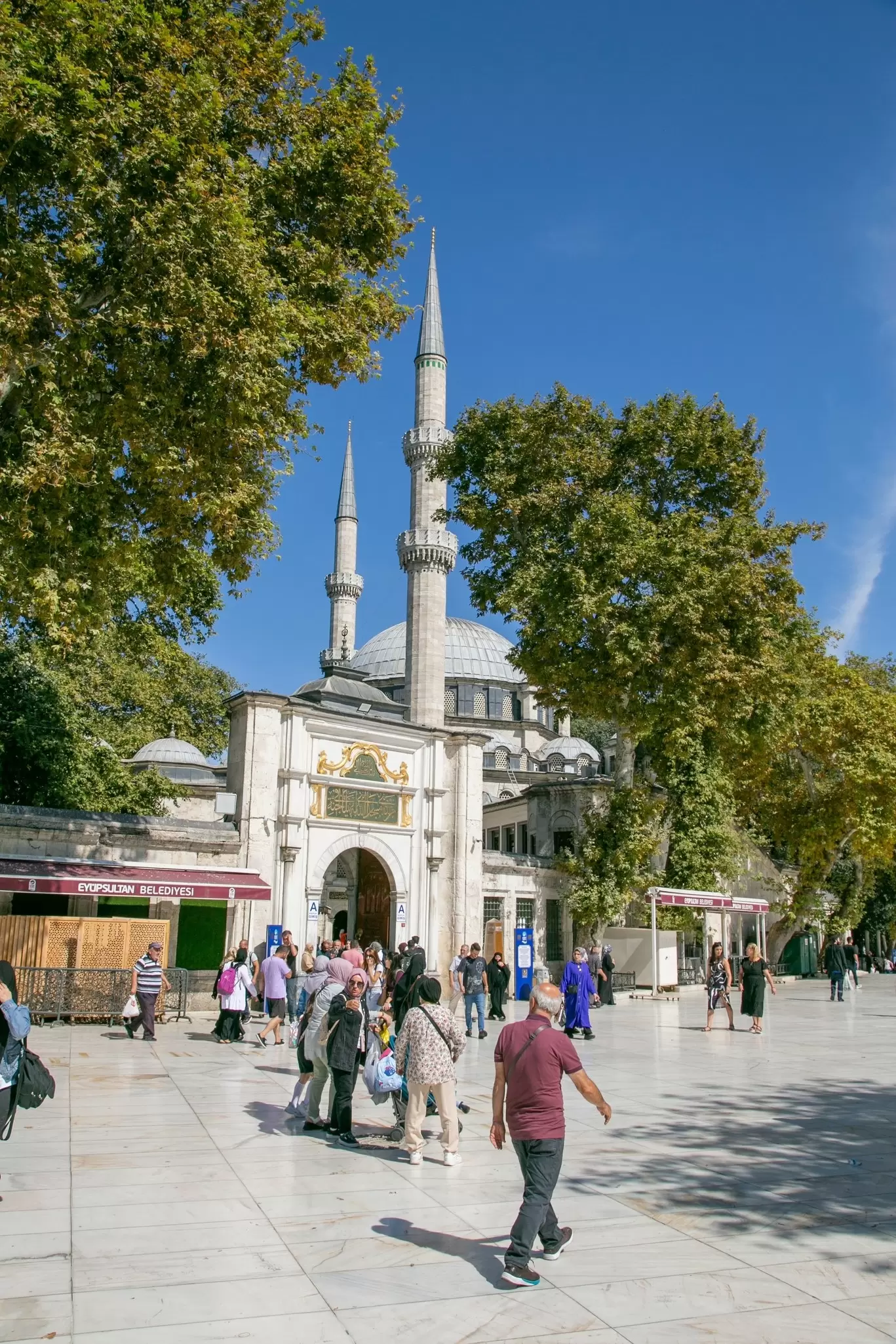 Visitors cross a sunlit marble plaza before the domed Eyup Sultan Mosque with its twin slender minarets, Istanbul.