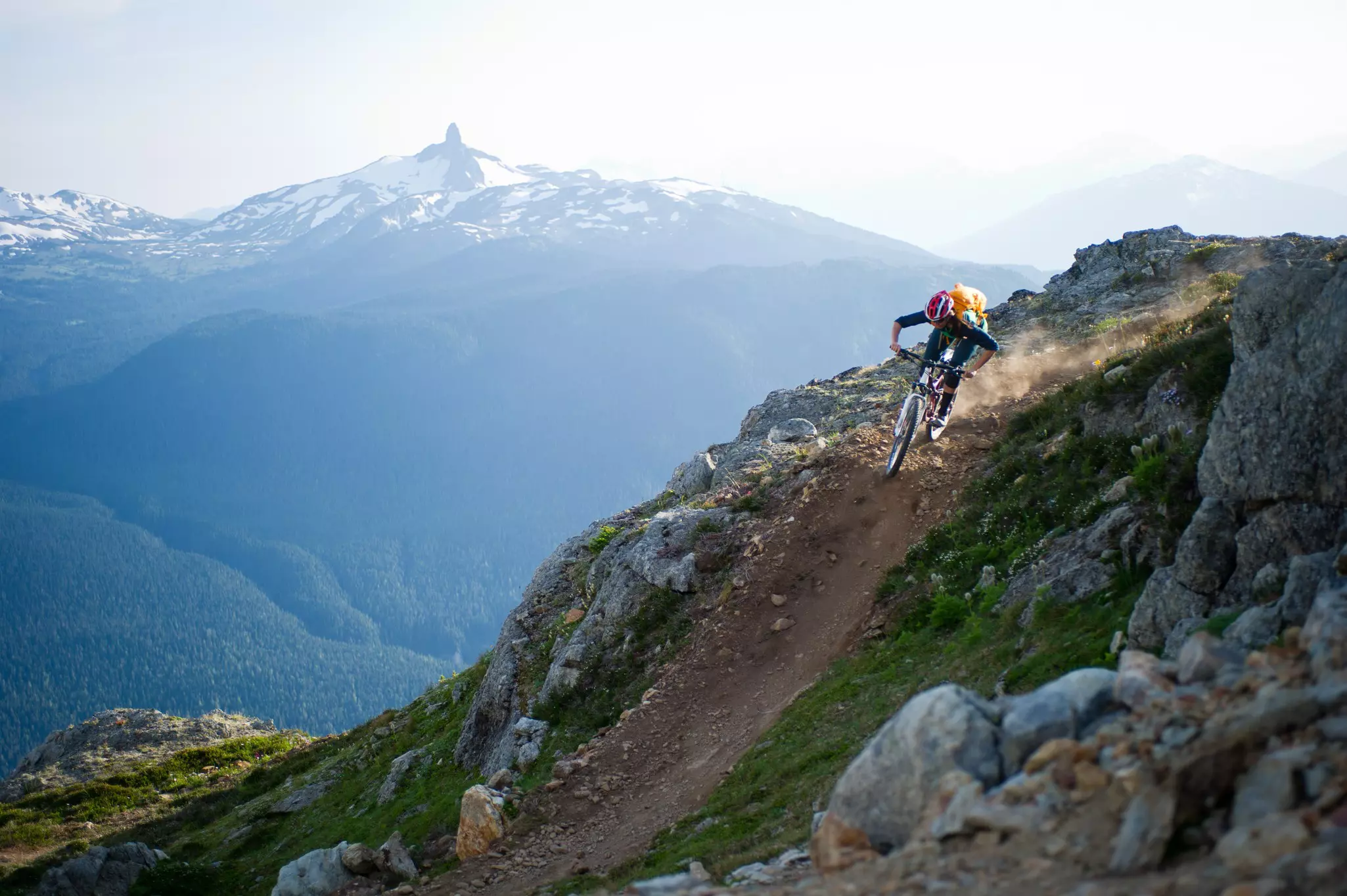 A mountain biker rides downhill on a dirt track in Canada; a snowcapped peak is in the background.