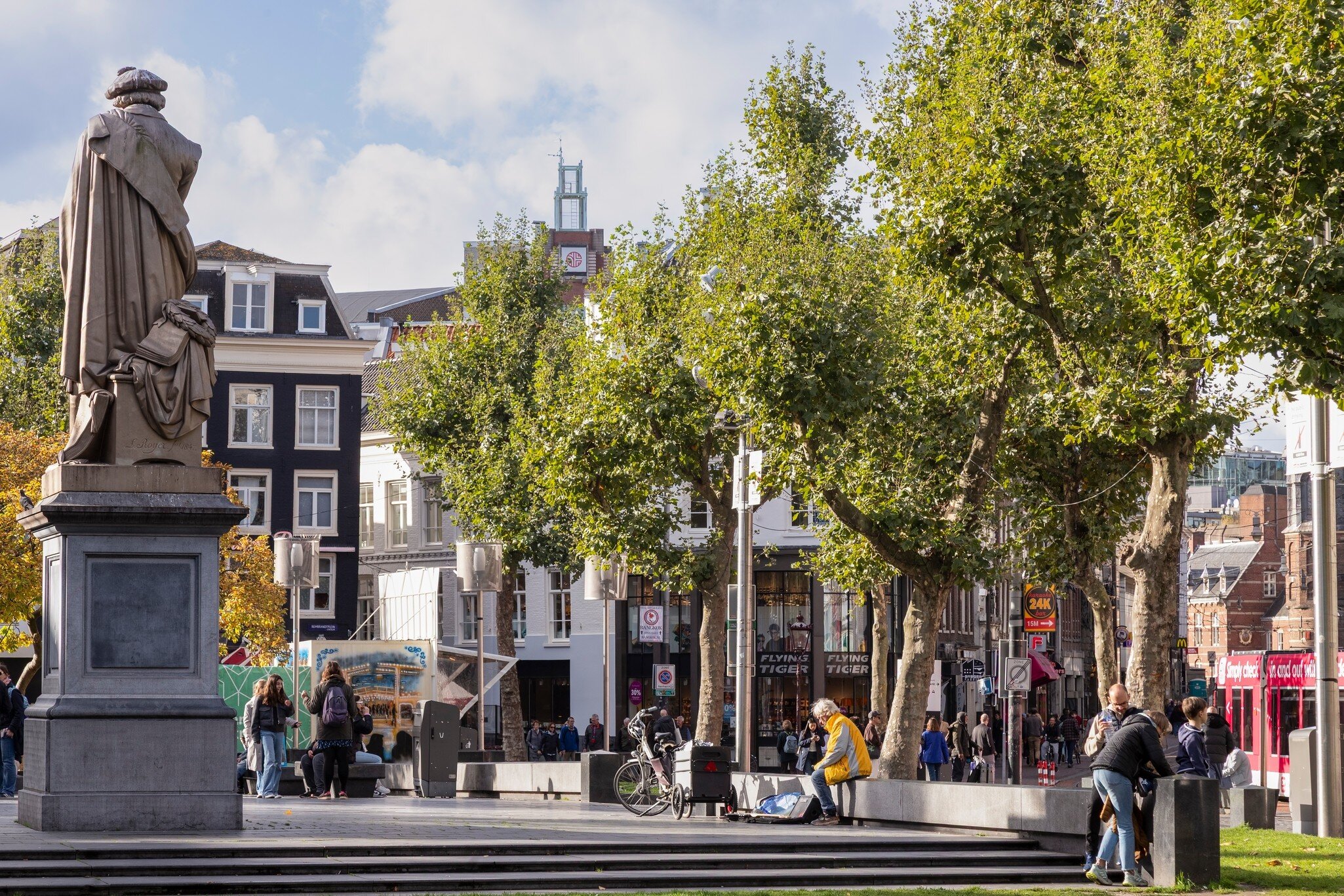 People enjoying the sunny autumn weather at the famous Rembrandtplein in Amsterdam, with a statue in the foreground.
