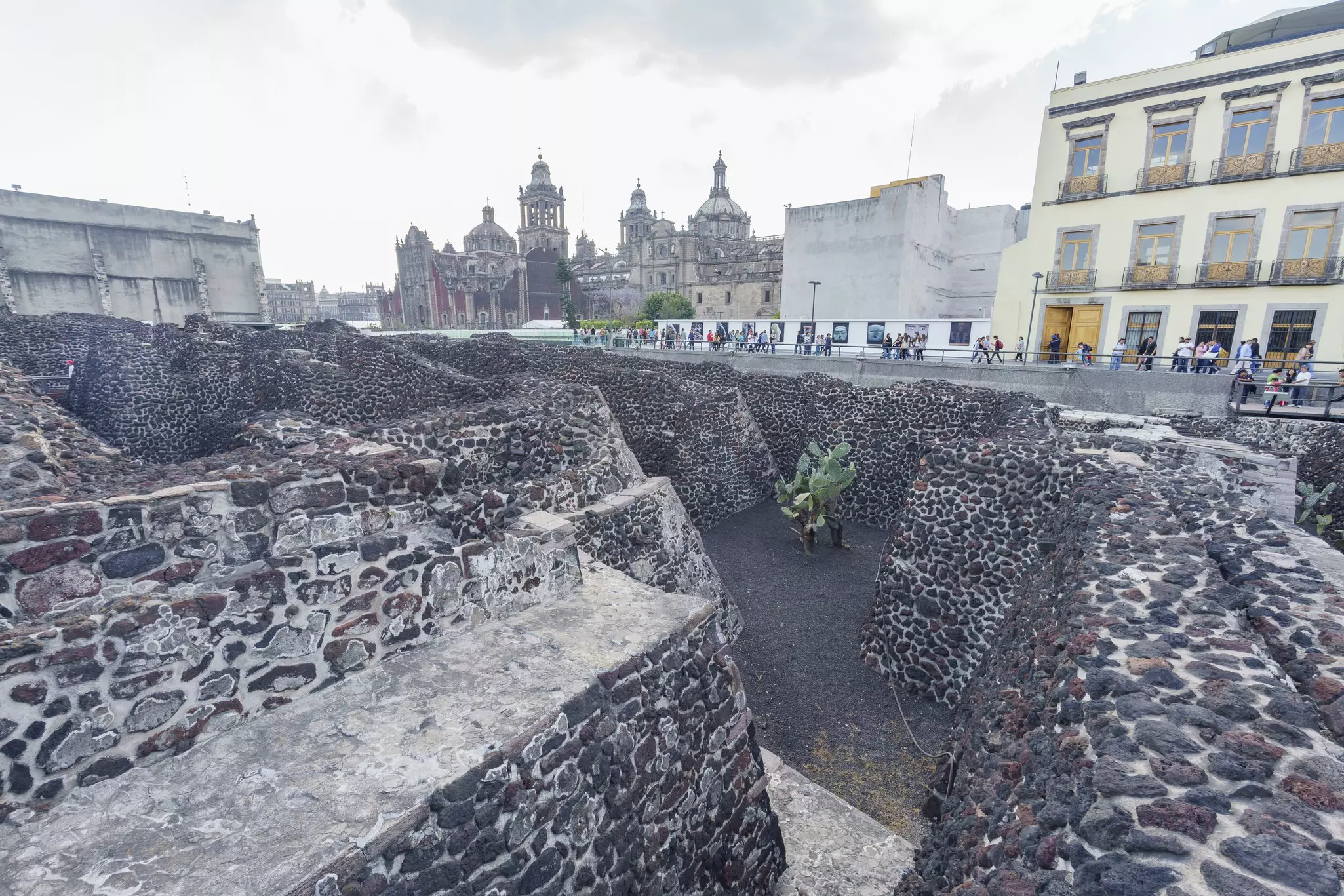 Mazelike brick stuctures in front of several buildings with people on a walkway between the two