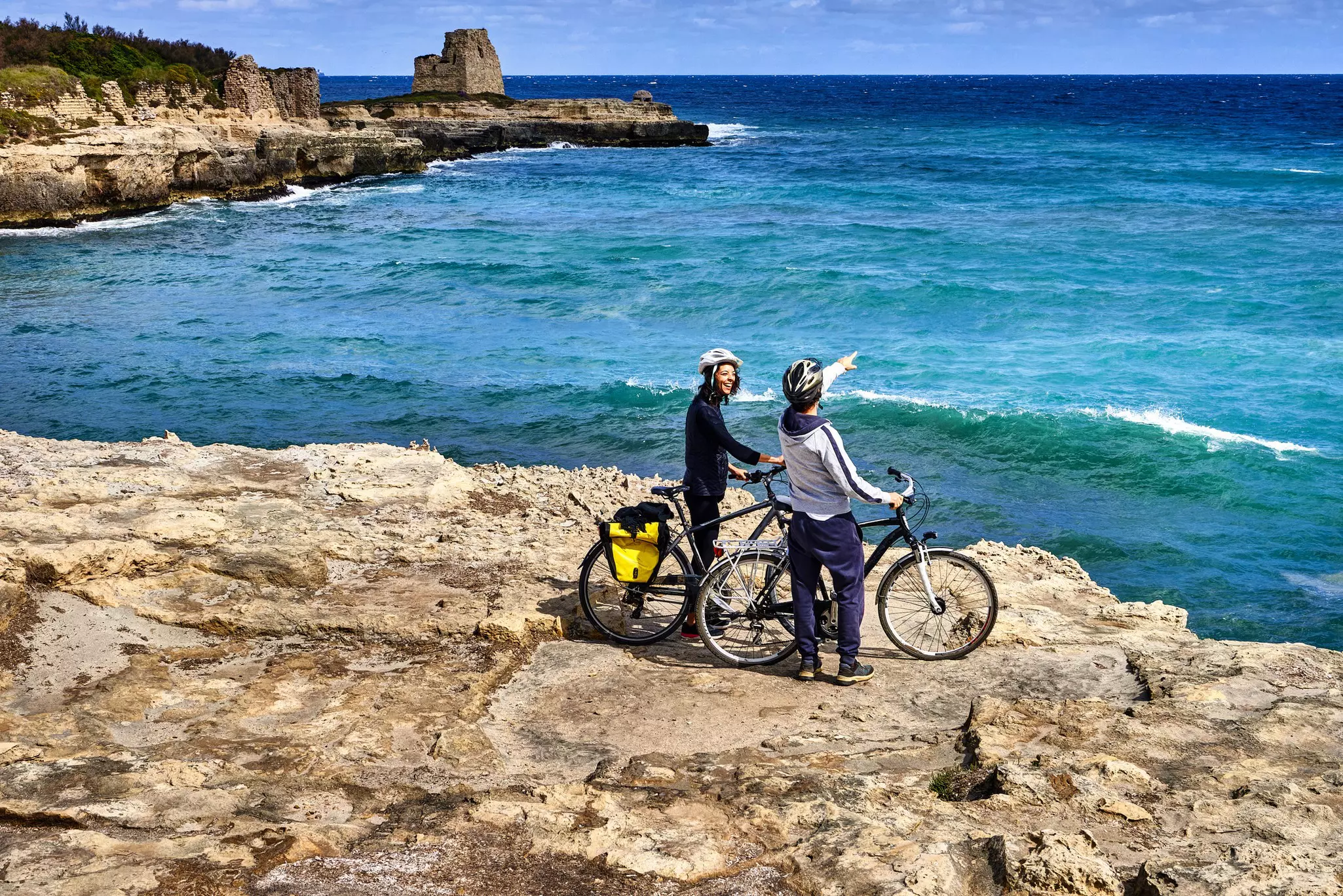 Two people on bicycles pause on a clifftop in Italy, looking out at the blue sea and rocky coastline