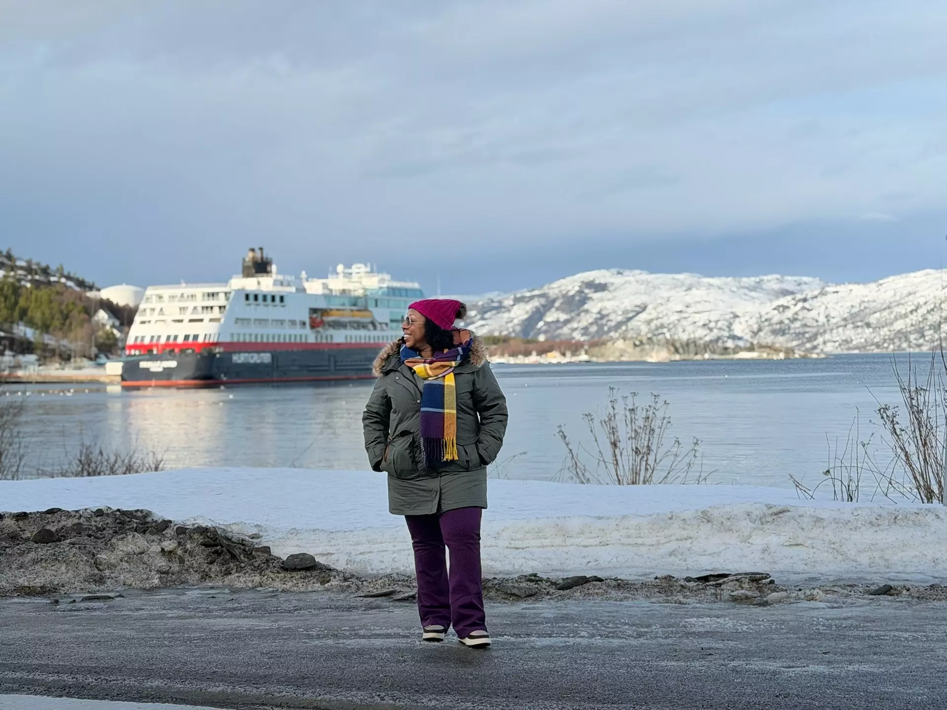 A woman in a heavy winter coat and colorful scarf stands by the shore with a large white ship in the background.