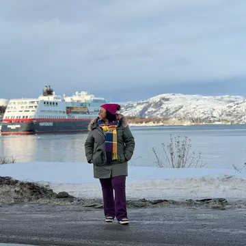 A woman in a heavy winter coat and colorful scarf stands by the shore with a large white ship in the background.