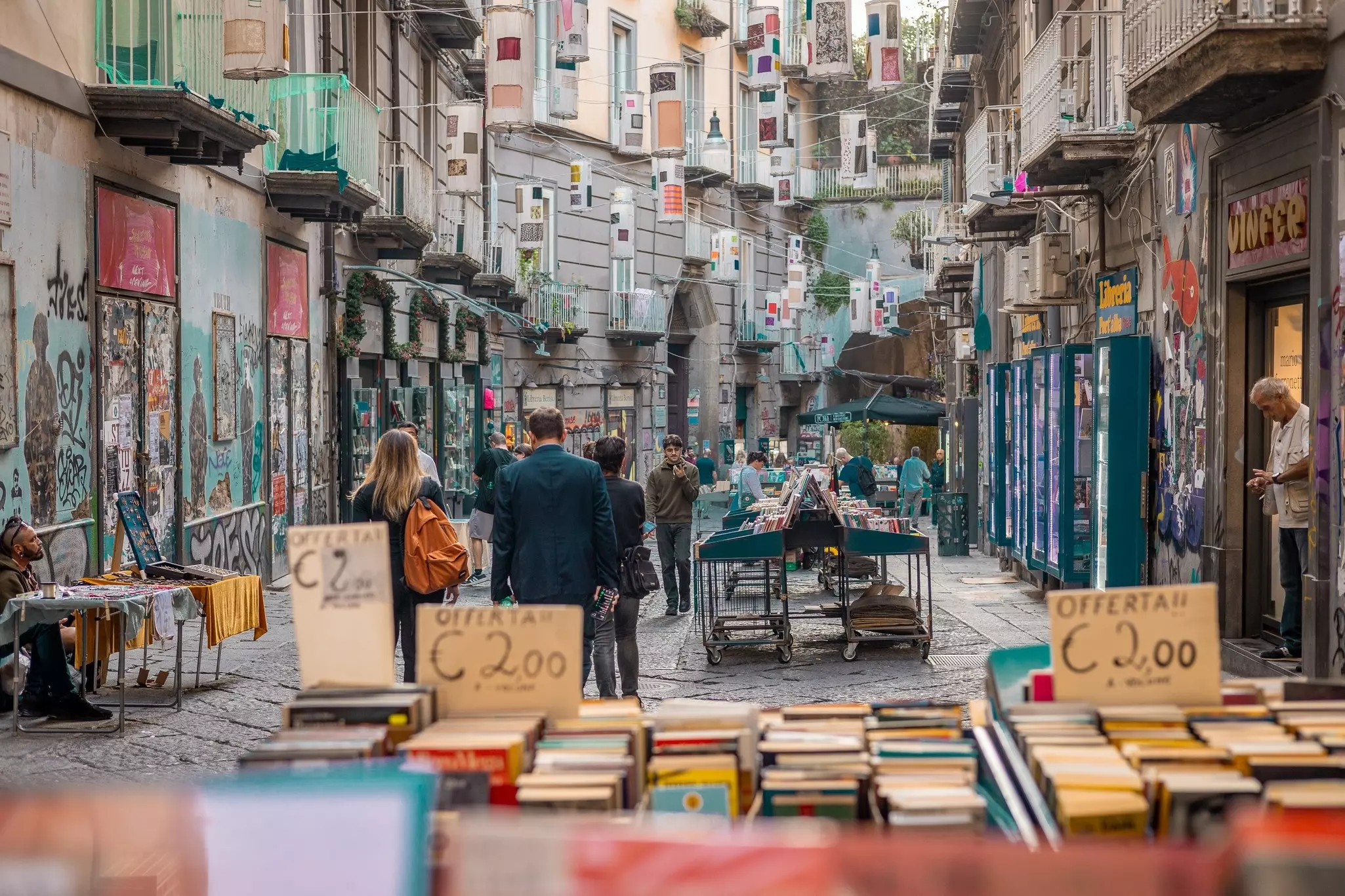 Pedestrians walk along a street lined with art and graffiti. Market stalls selling books and trickets stand in the street.
