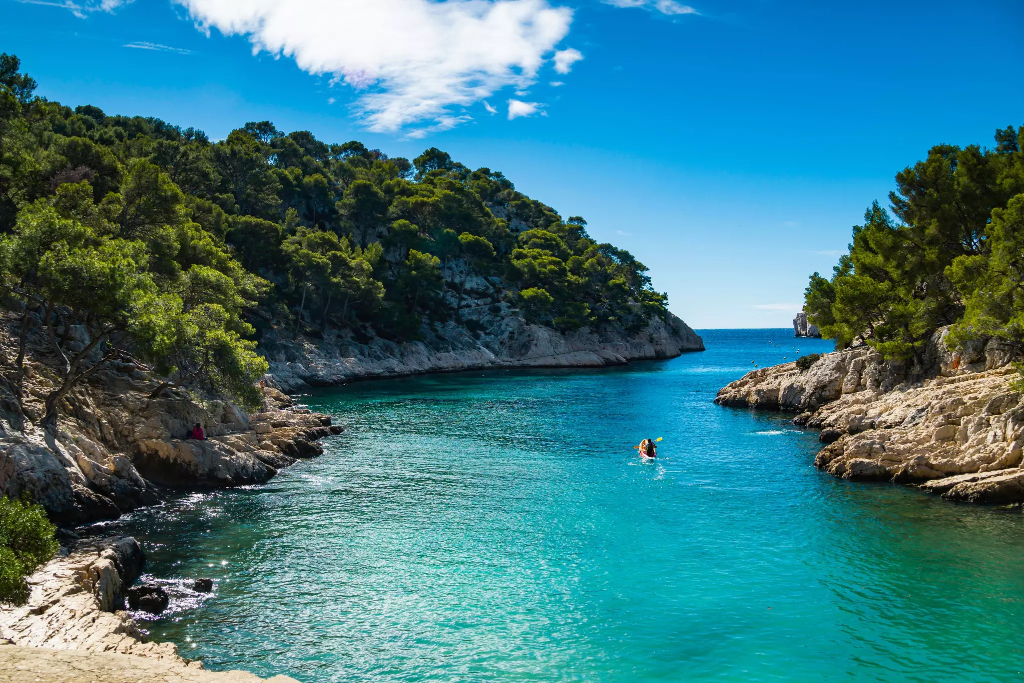 One person kayak on blue water in craggy coastline with trees, sky and clouds up ahead