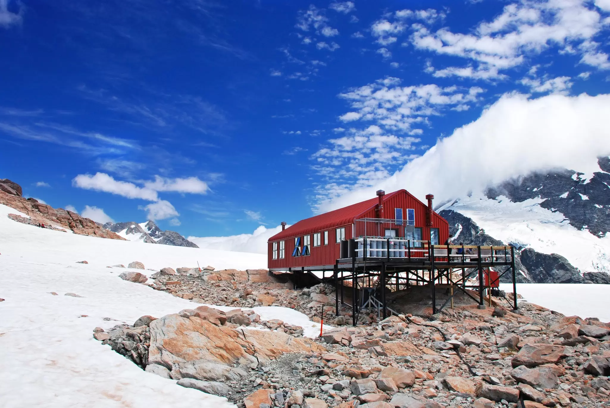 A large red metal hut surrounded by snow-covered hills and rocky terrain