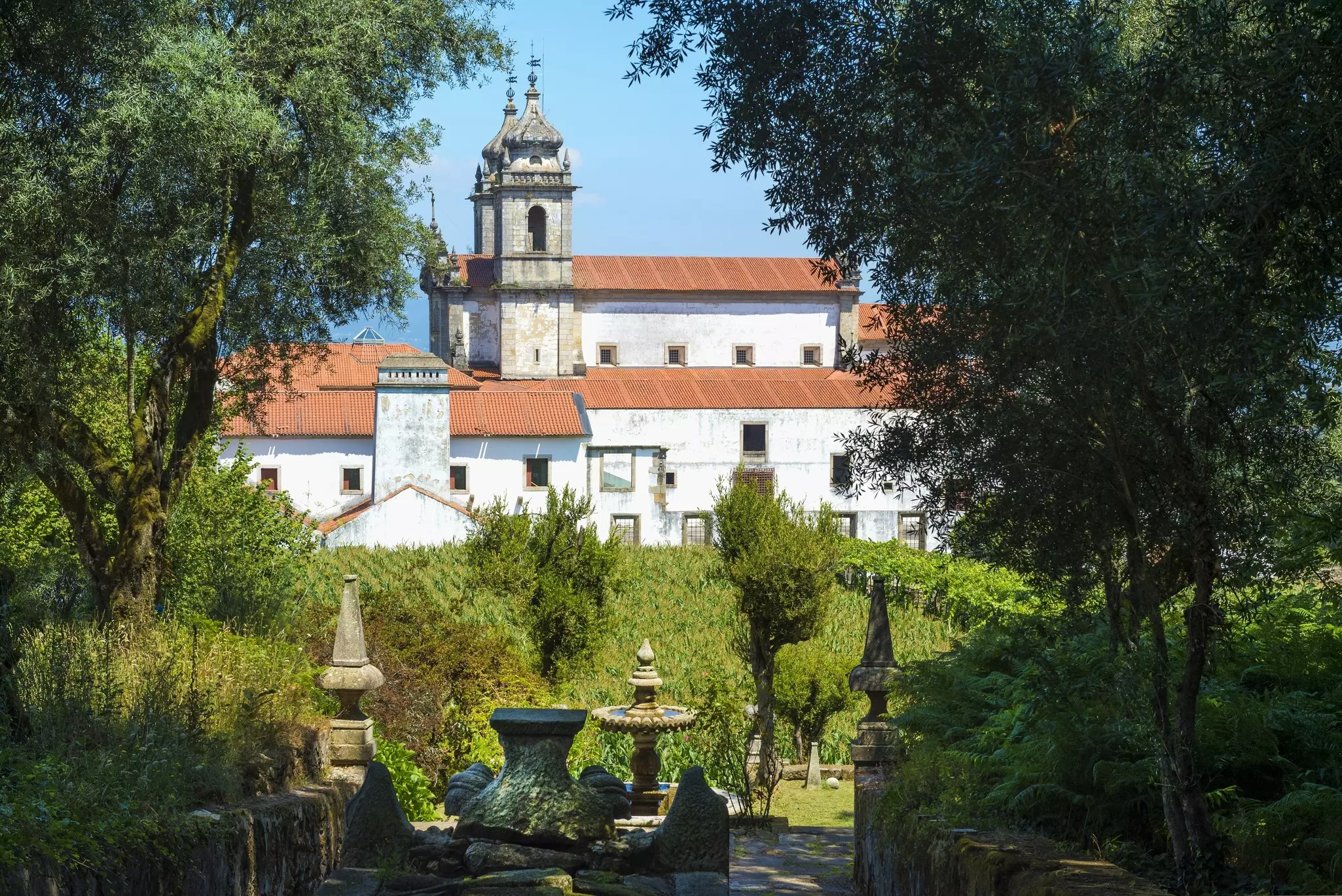 A large church with red-tiled roofs and white walls is seen through trees; there is a garden with some sculptures in the foreground.