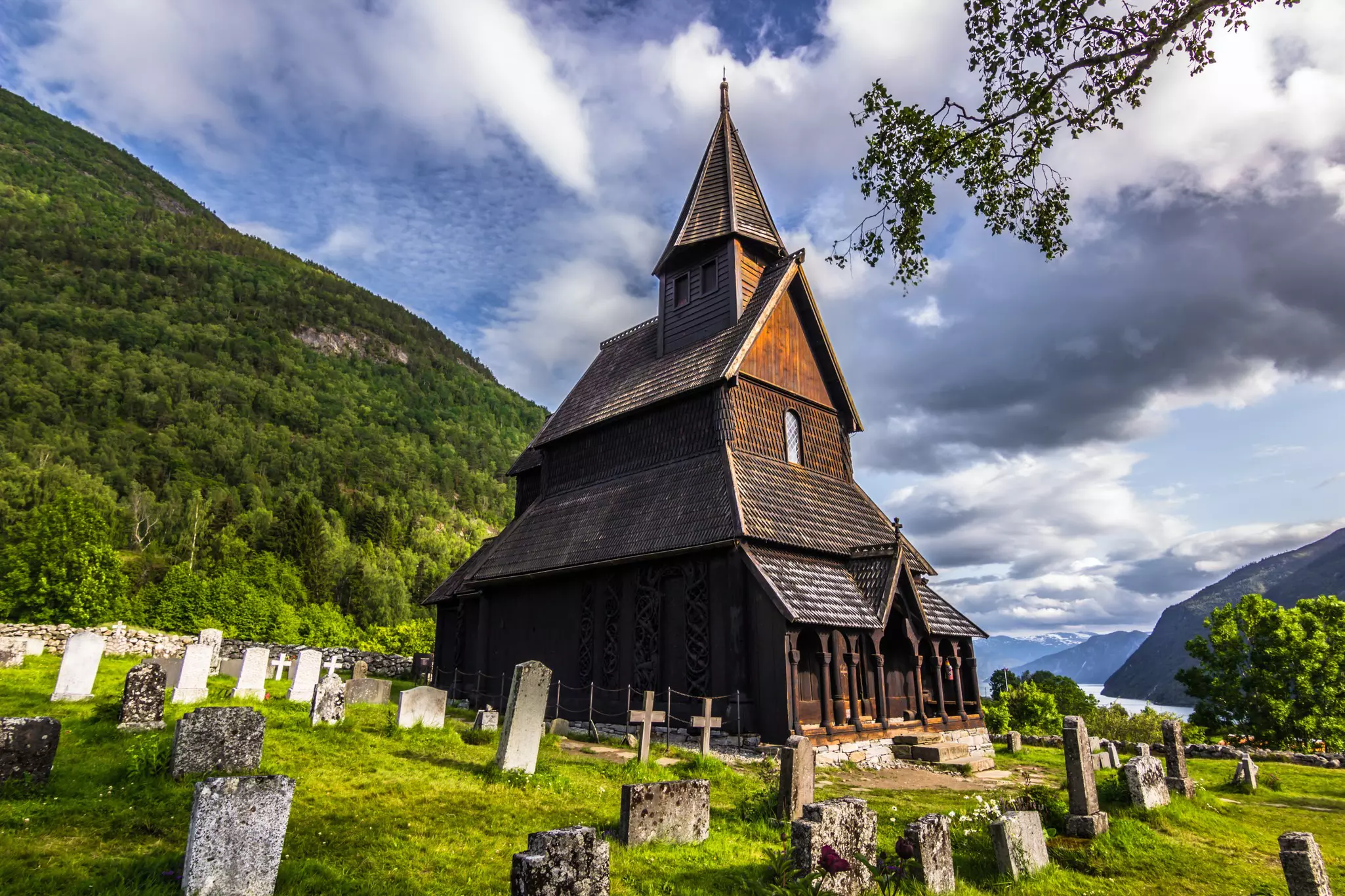 A view of the cemetery in front of a historic church made of wood planks. Verdant hills and a fjord are seen in the distance.