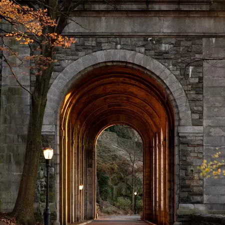 An arched entry surrounded by stonework in a city park with a hint of sun coming through autumnal-colored tree.