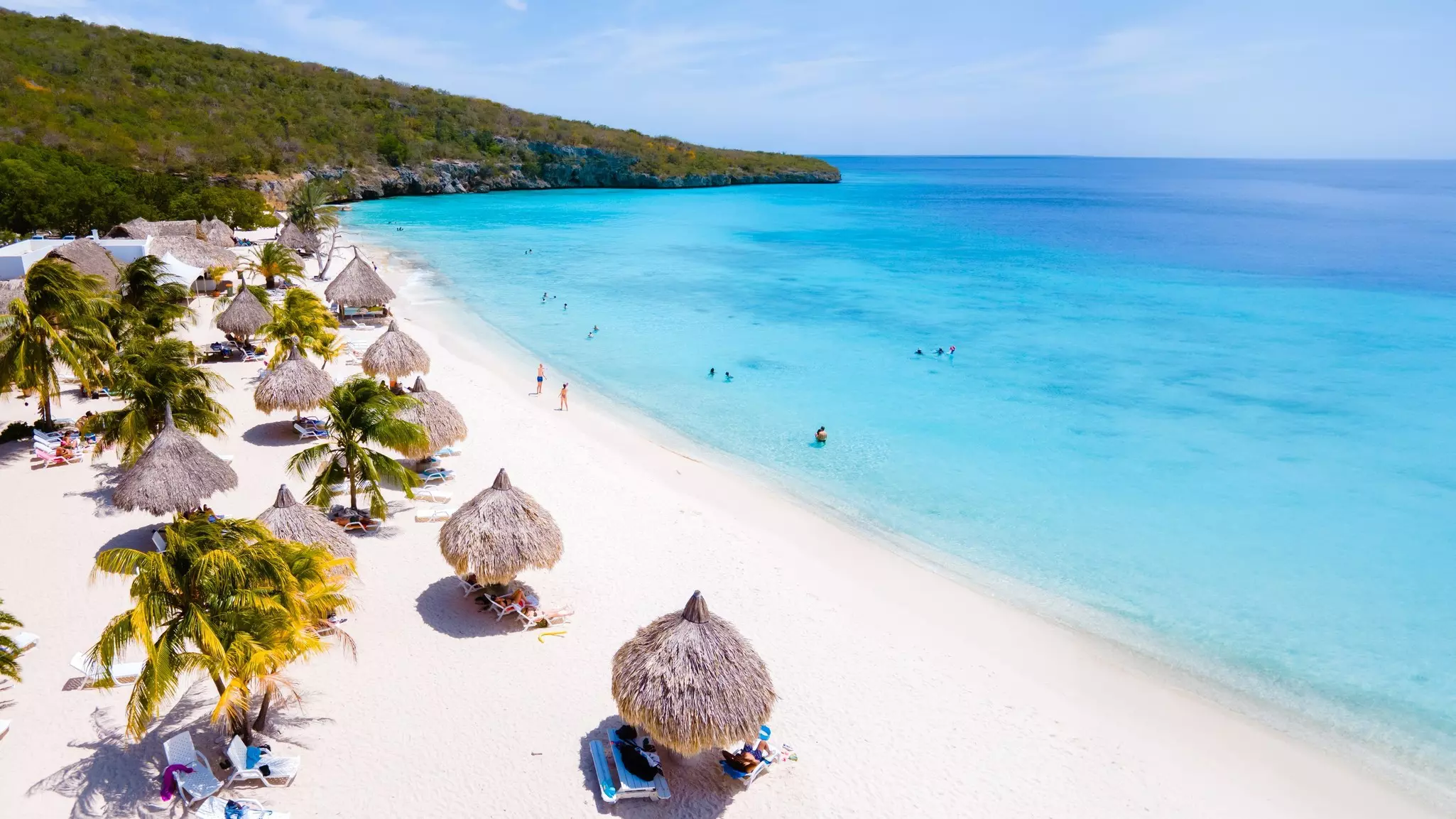 A lovely stretch of bright white sand lined with thatched shades. People play on the beach and splash in the turquoise ocean.