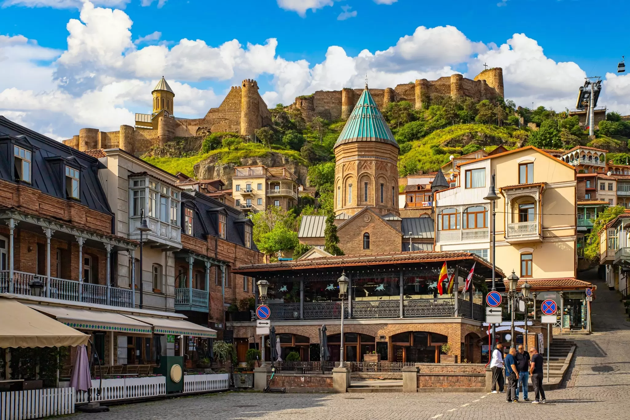 Tbilisi old town with wooden-balconied houses and a turquoise-domed church below the ancient Narikala fortress and a cable car