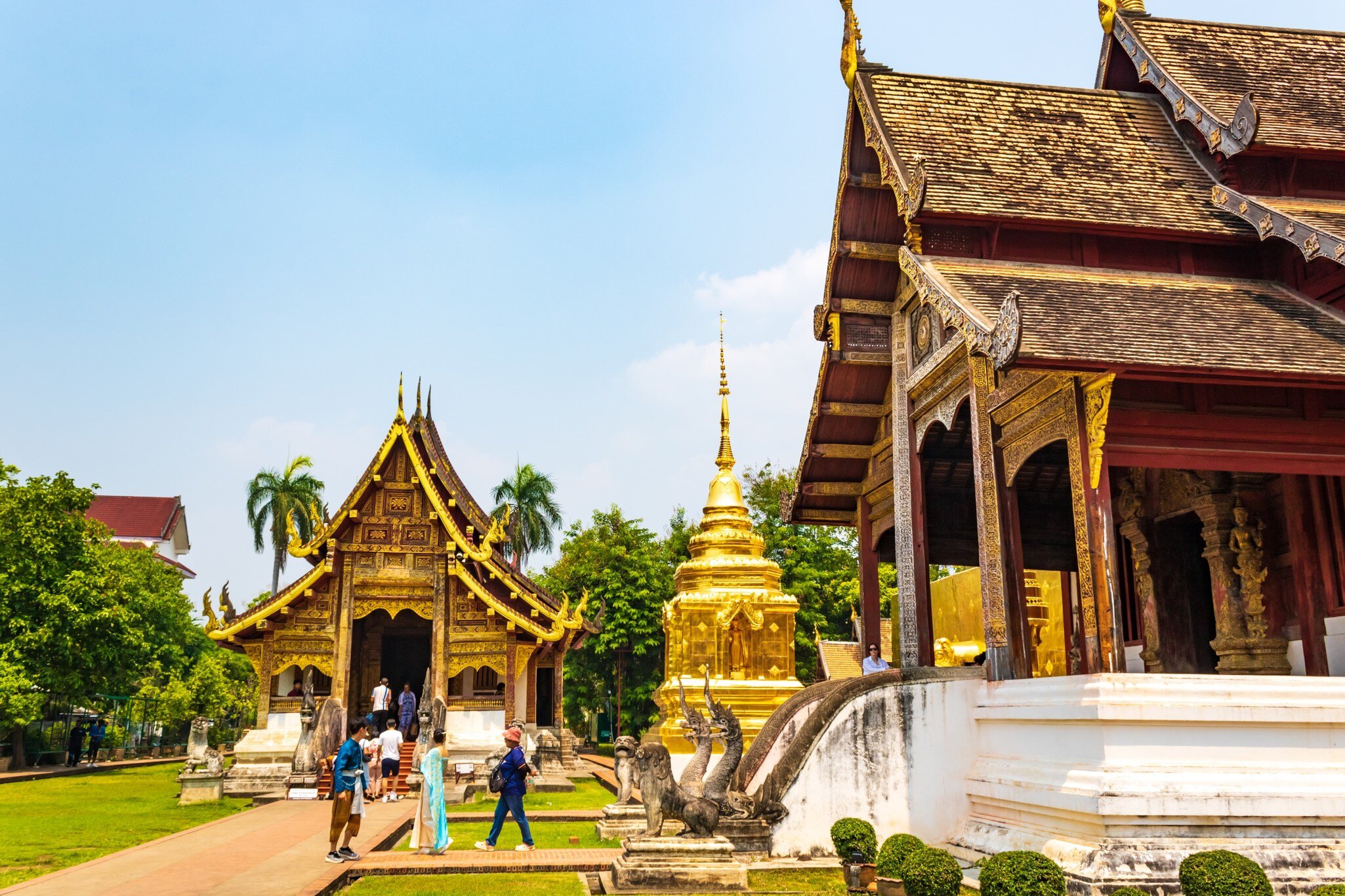 People walk around the grounds of a temple complex with golden features on its buildings and shrines.