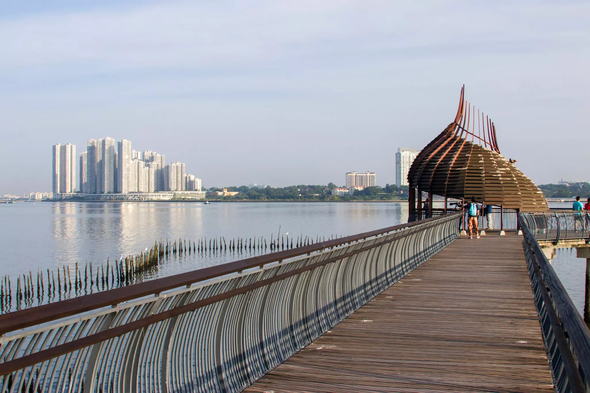 A walkway over water leads to a thatched-roof observation point. Apartment buildings are seen on the far shore.