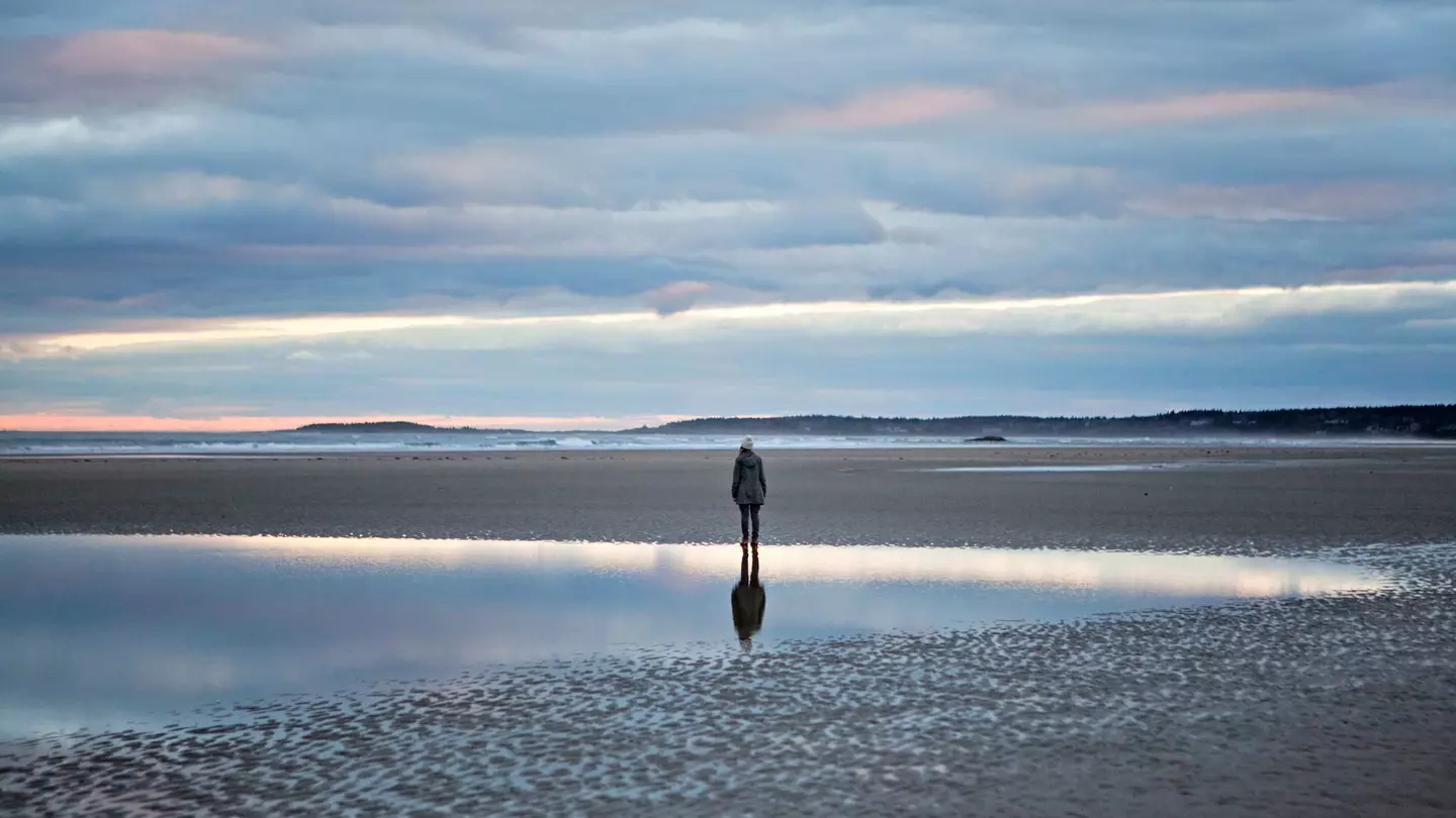 A woman stands reflected in a tidal pool on the beach in Maine
