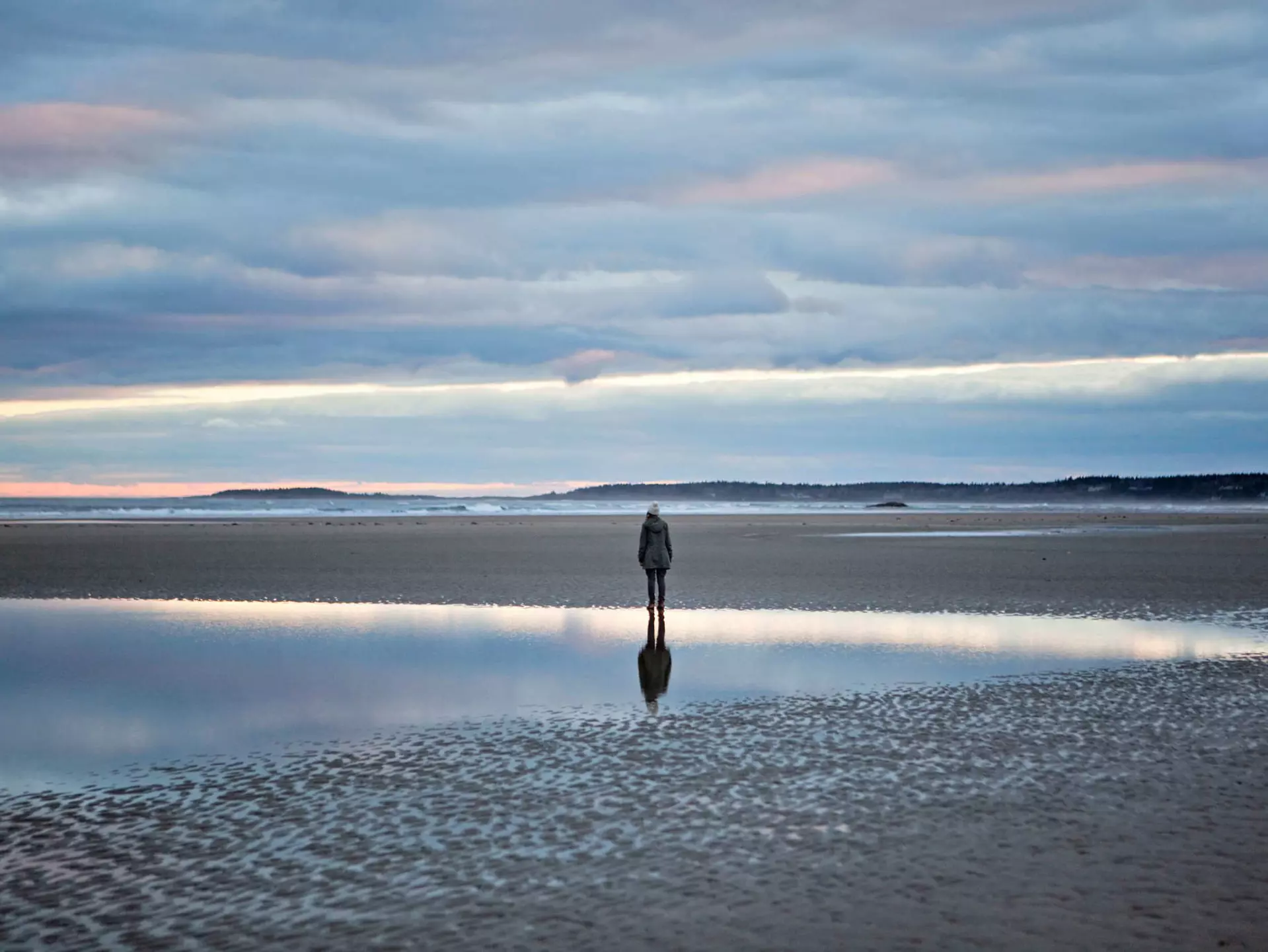 A woman stands reflected in a tidal pool on the beach in Maine