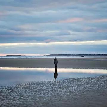 A woman stands reflected in a tidal pool on the beach in Maine
