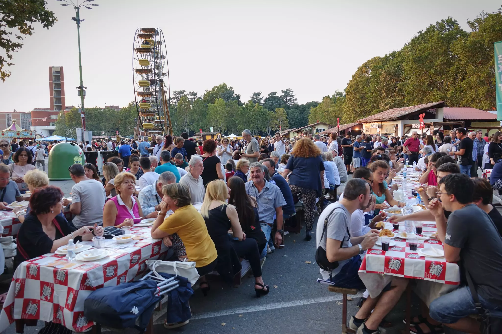 People eat together at communal tables with tableclothes in a town square during a festival.