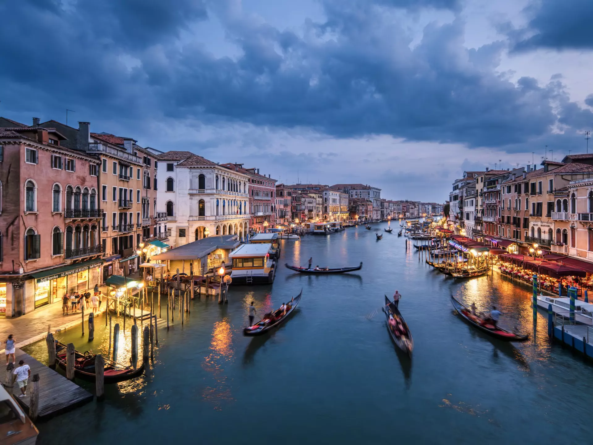 A canal at night. Narrow boats glide down the center as people dine at restaurants around the edge.