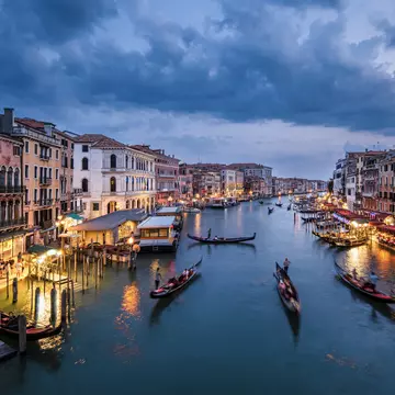 A canal at night. Narrow boats glide down the center as people dine at restaurants around the edge.