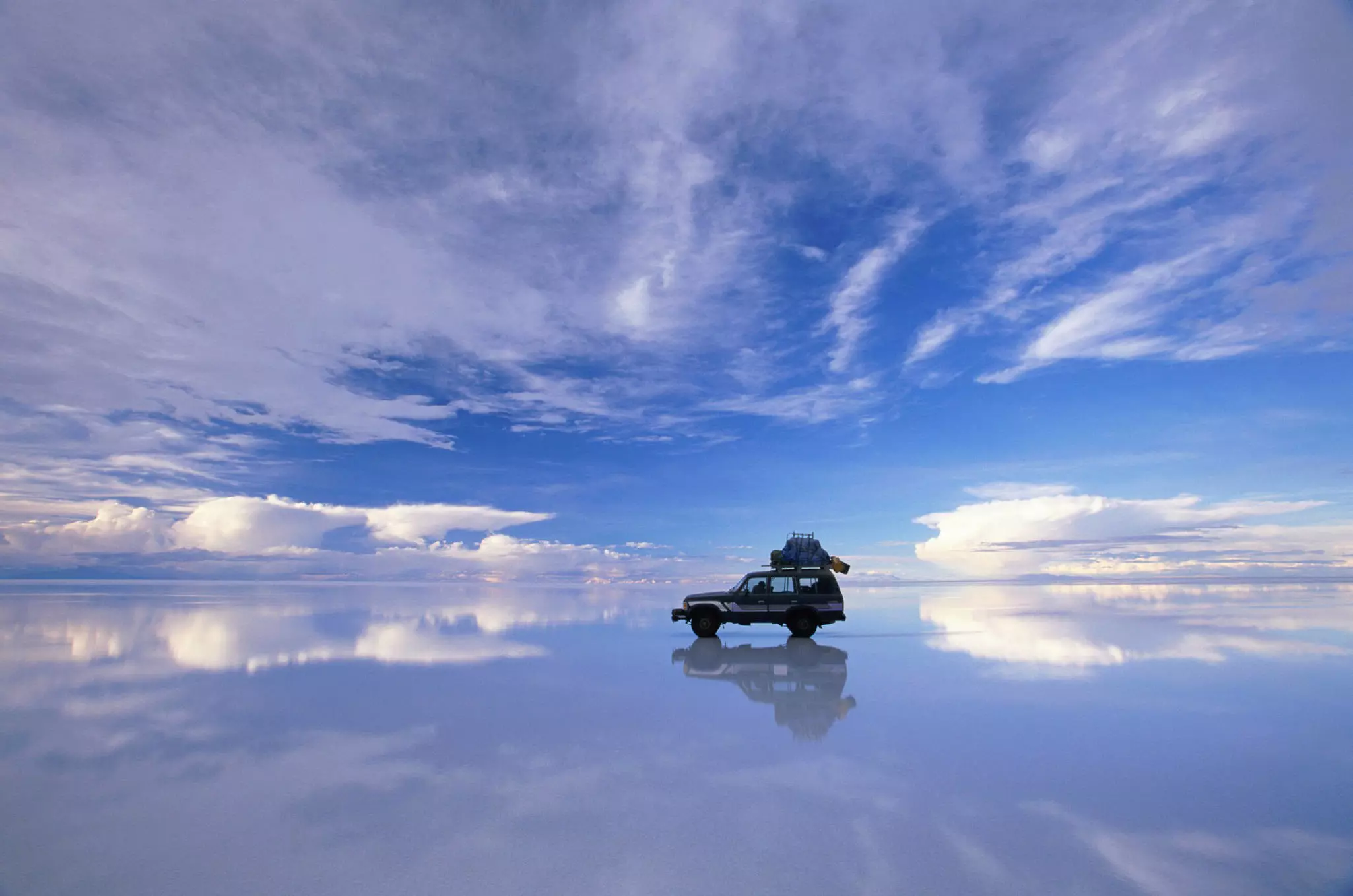 A four-wheel drive reflected in salt flats appears to float between the water and the sky