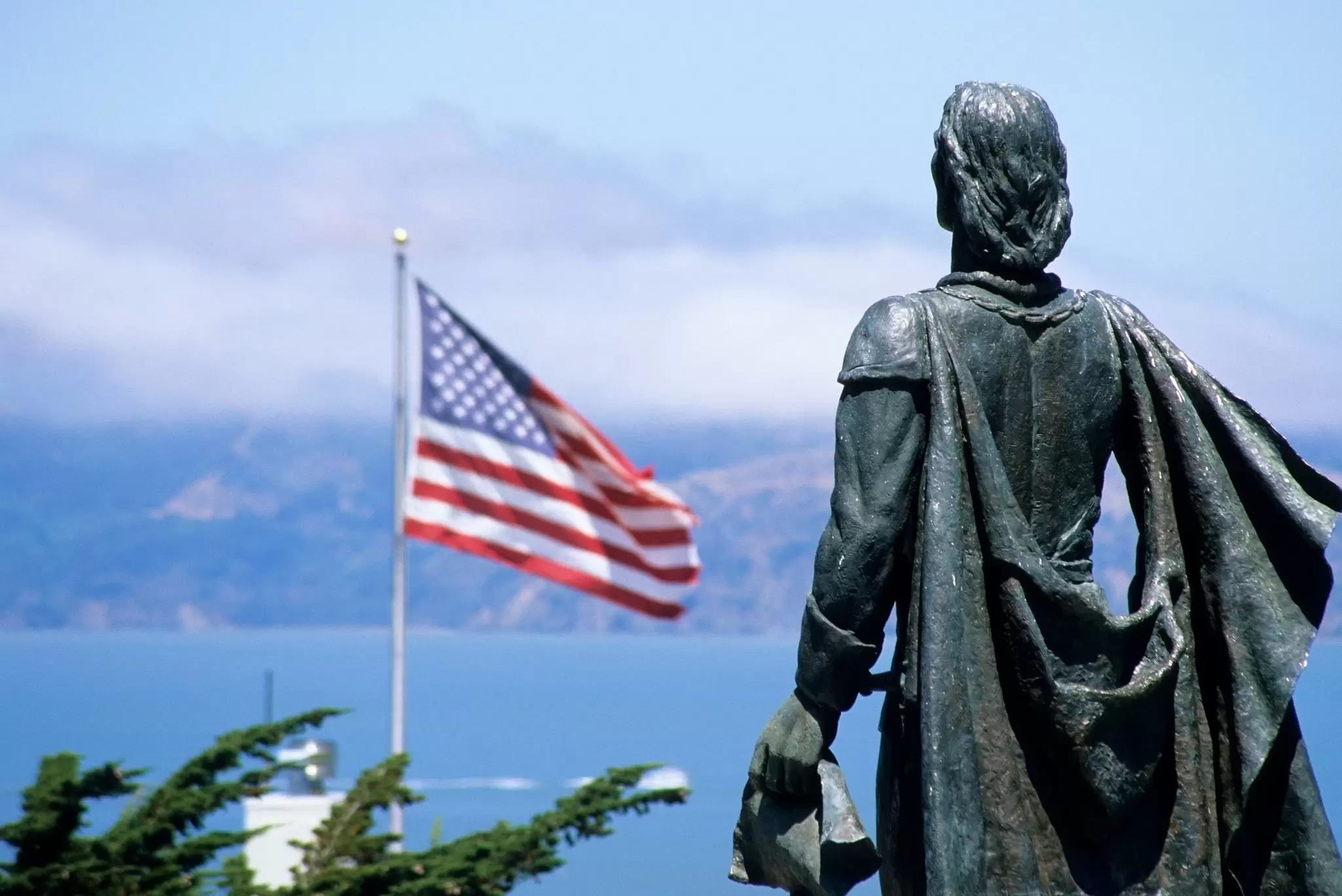 A bronze statue of a cloaked figure looking out over the sea toward a waving American flag
