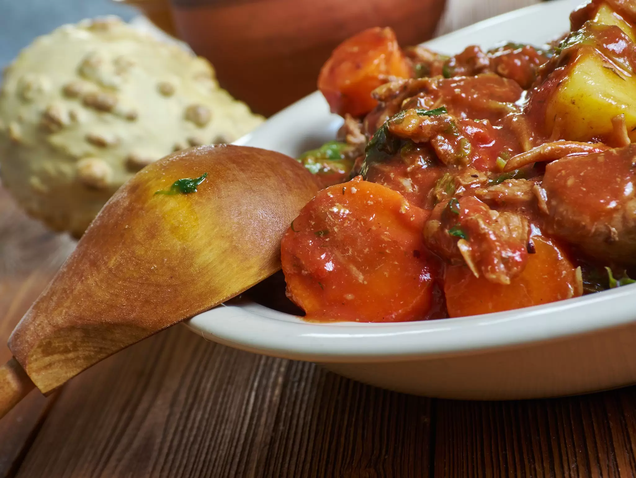 Close-up of a meat and vegetable curry in a white bowl beside a piece of naan and a bread roll.