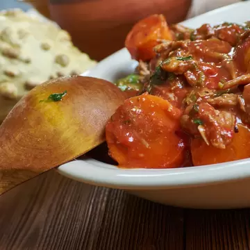 Close-up of a meat and vegetable curry in a white bowl beside a piece of naan and a bread roll.