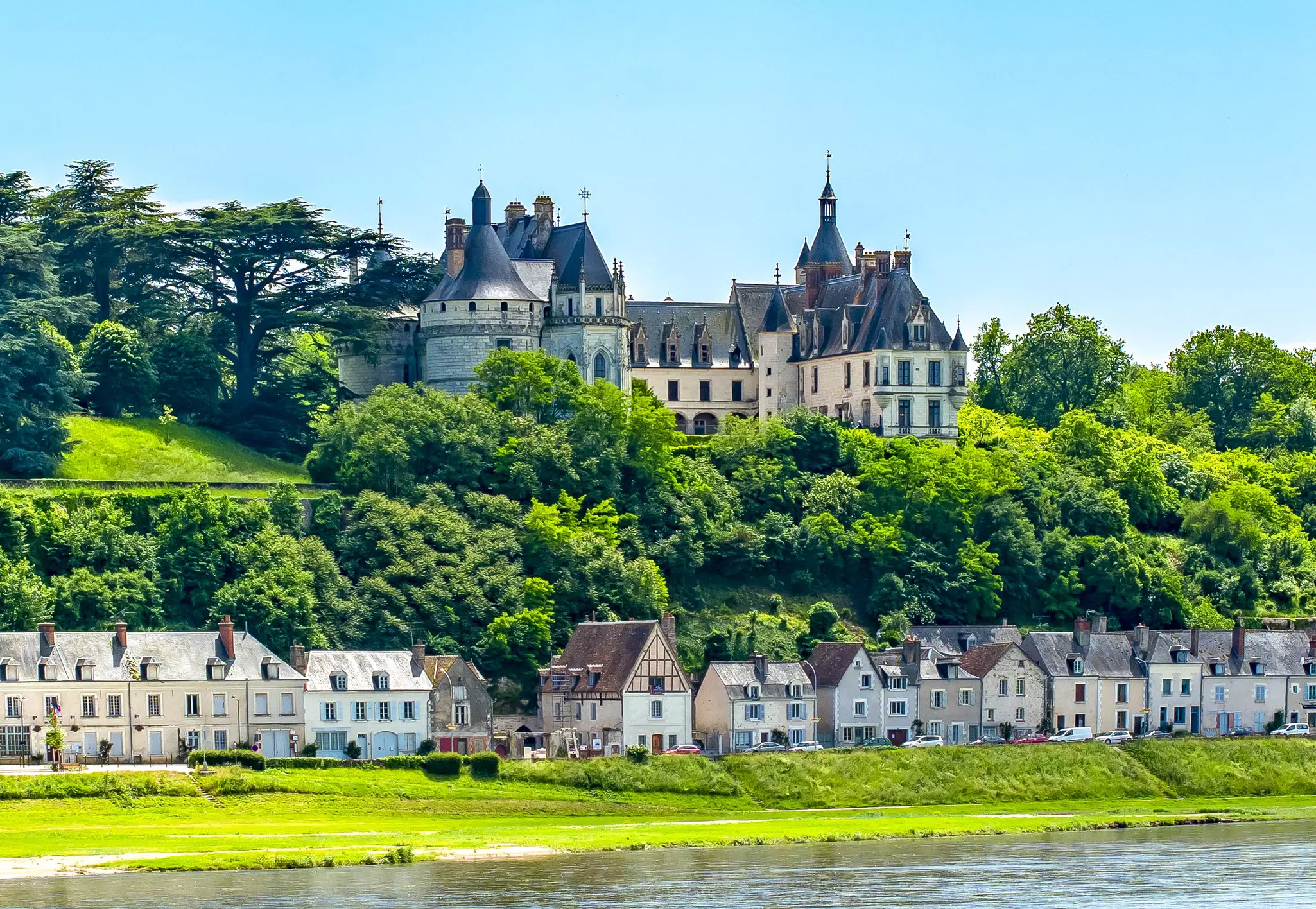 Houses line the green banks of a riverside overlooked by a large chateau.