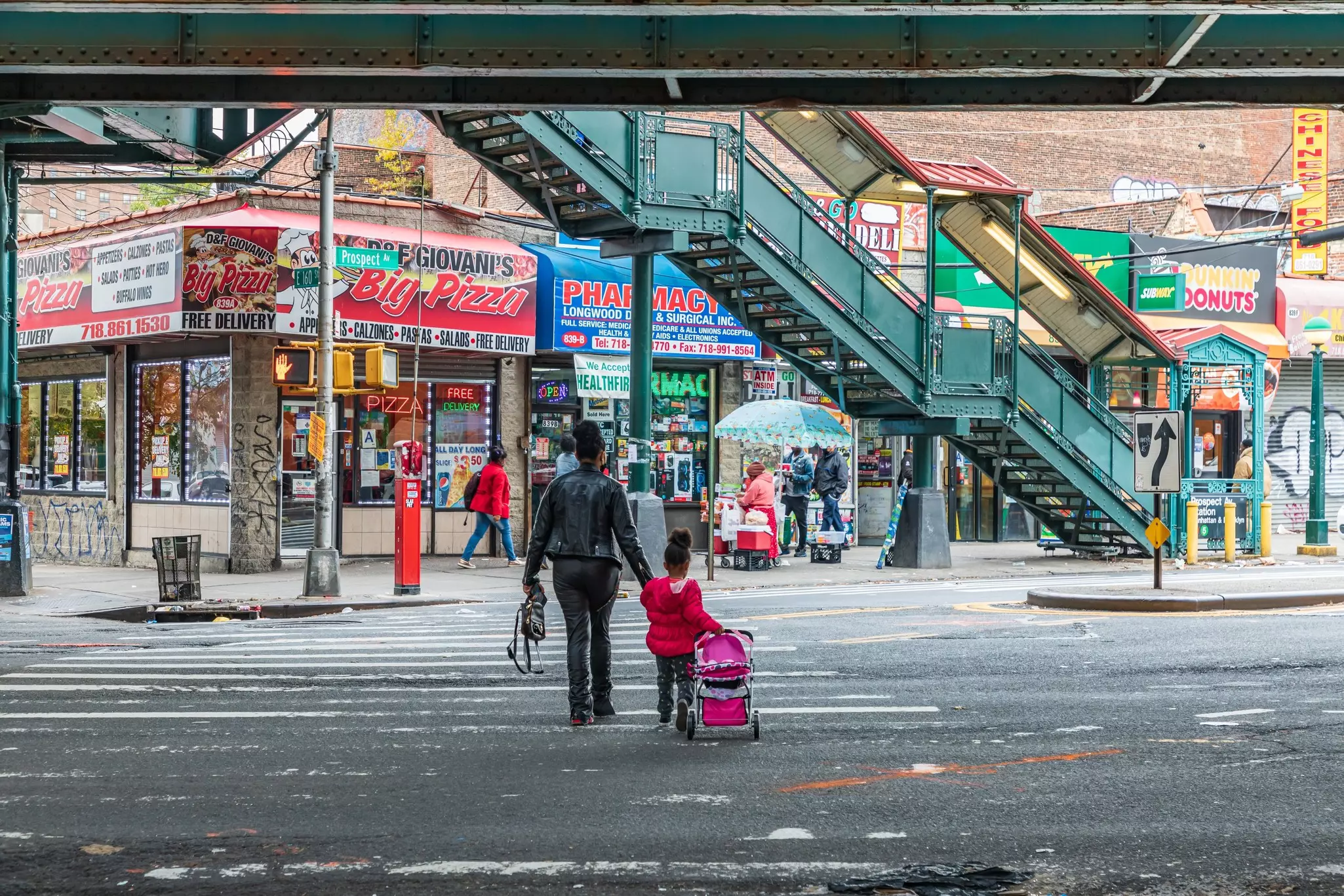 A woman and a child cross the street toward stores and the entrance to the overhead metro station.