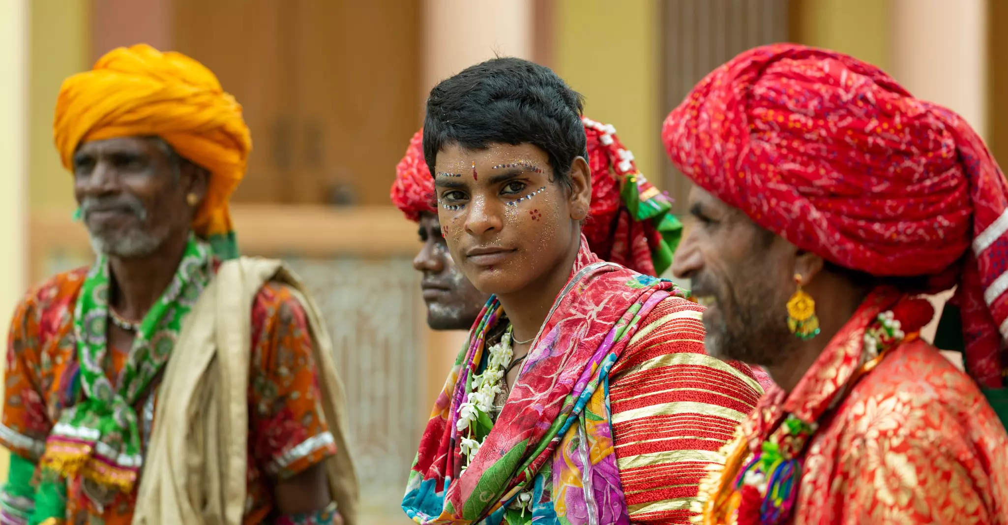 Four men wearing clothes with rich, vibrant colors. Some wear turbans, earrings and face paint.