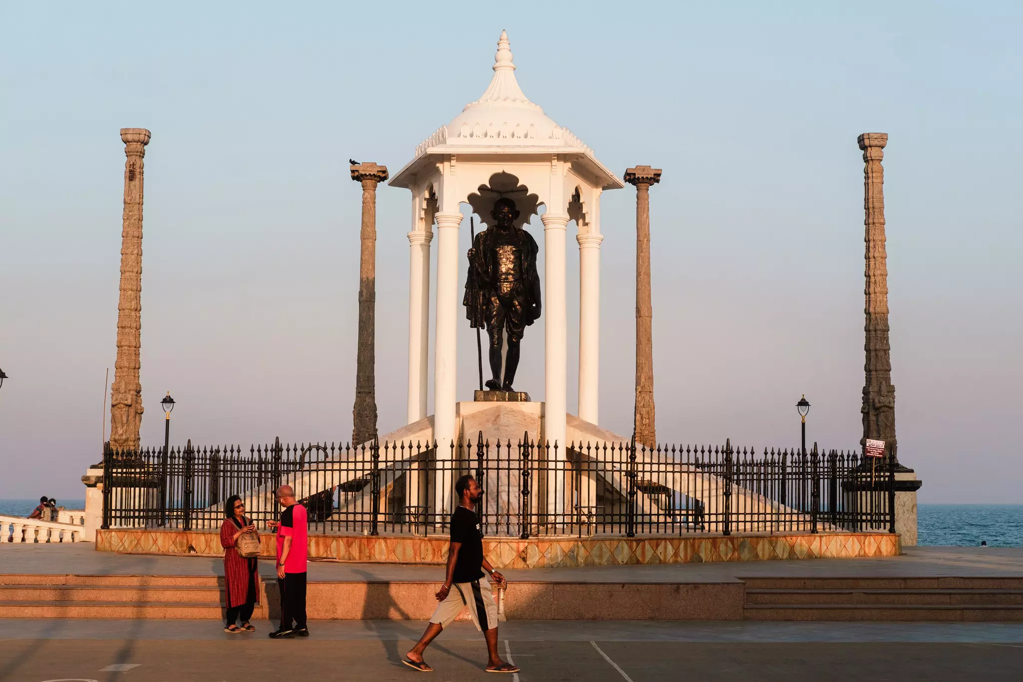 People walk past a full-length statue of a person under a white enclosure; the sea is on the other side of the path.