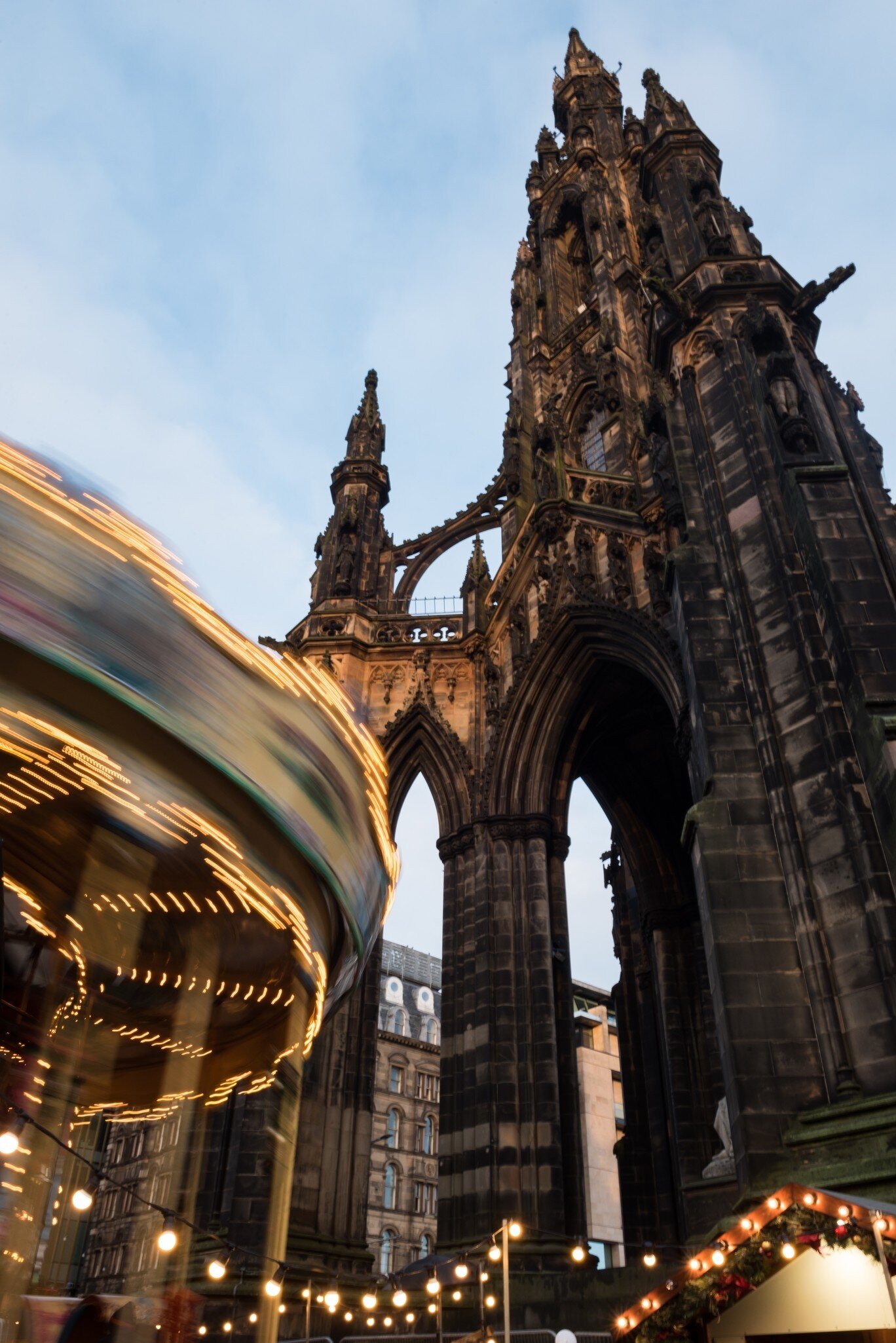 An illuminated vintage carousel spins next to the historical Walter Scott Monument during Christmas time in Princes Street Gardens under a blue sky with clouds.