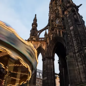 An illuminated vintage carousel spins next to the historical Walter Scott Monument during Christmas time in Princes Street Gardens under a blue sky with clouds.