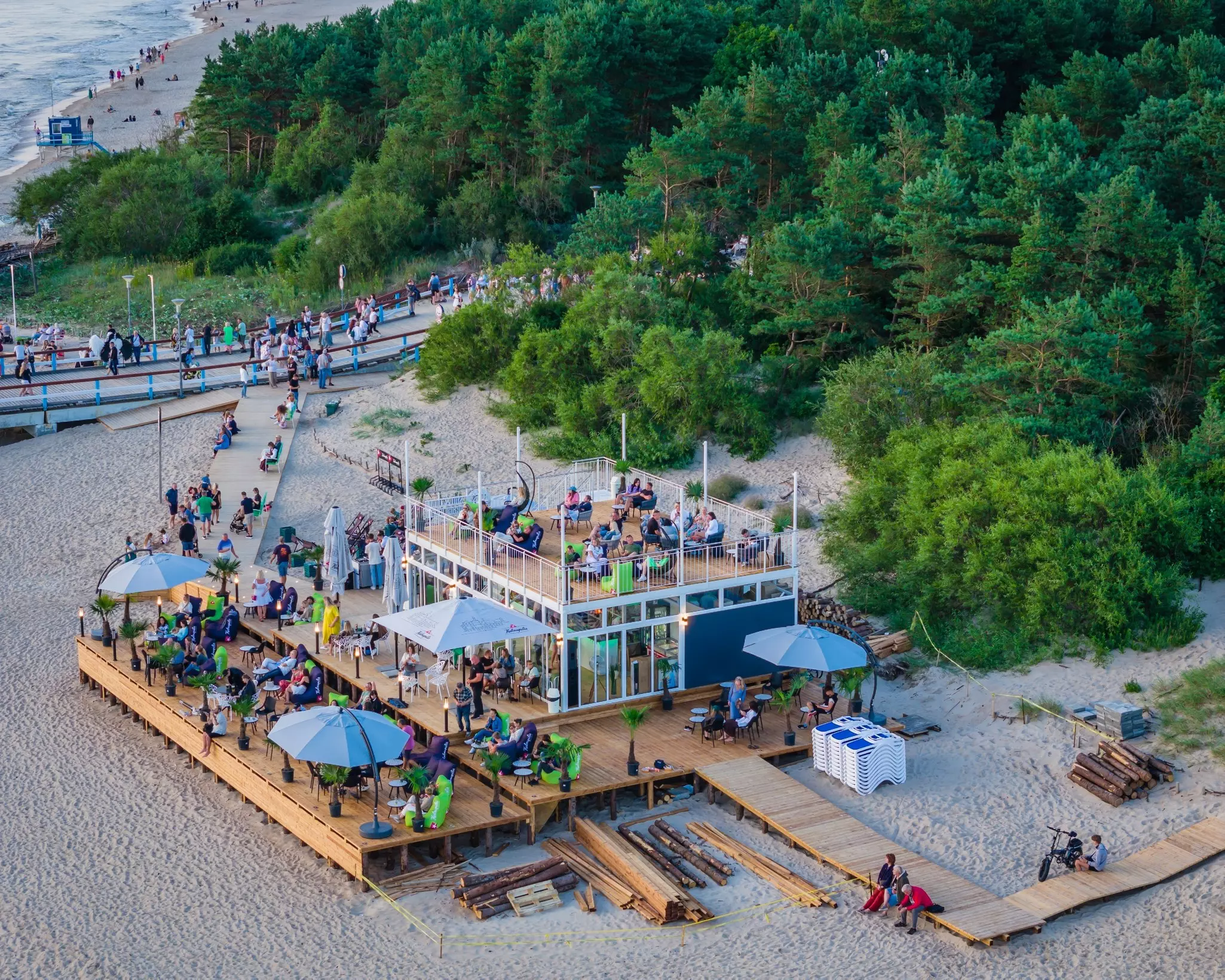 A beach bar with decking backed by woodland. Patrons sit on comfortable chairs.