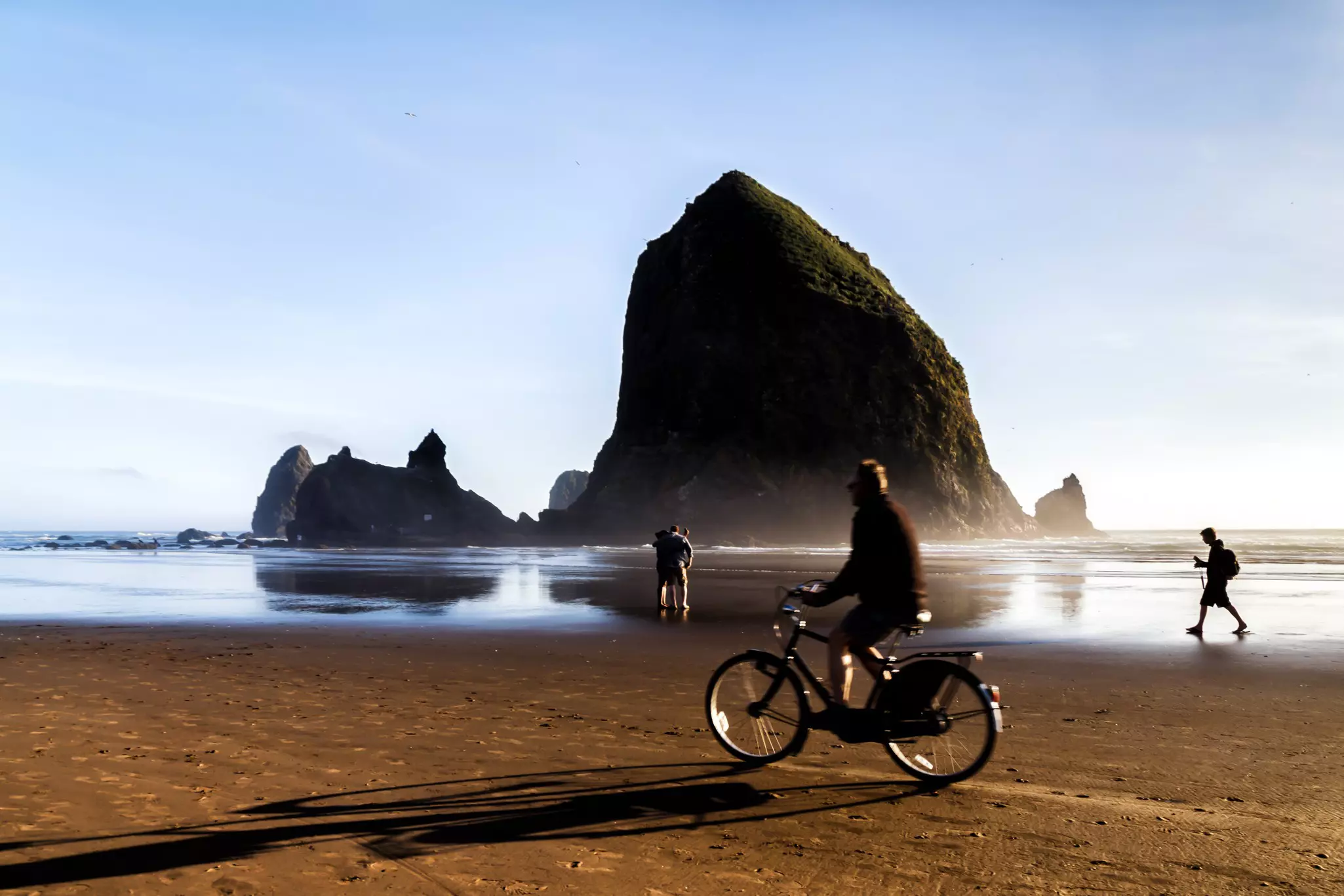 Haystack Rock at Cannon Beach, landmark of the Oregon Coast. Silhouettes of a couple making selfie, bicyclist passing by. Blurred movement. 