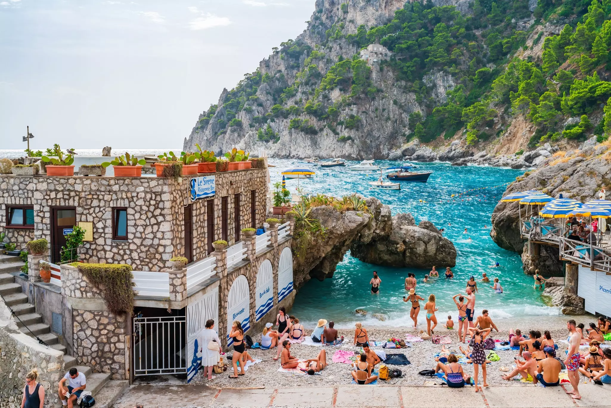 A group of people in swimsuits on a beach in Marina Piccola on Capri Island, Italy, with stone buildings on the left and the sea and rocky hill in the distance..
