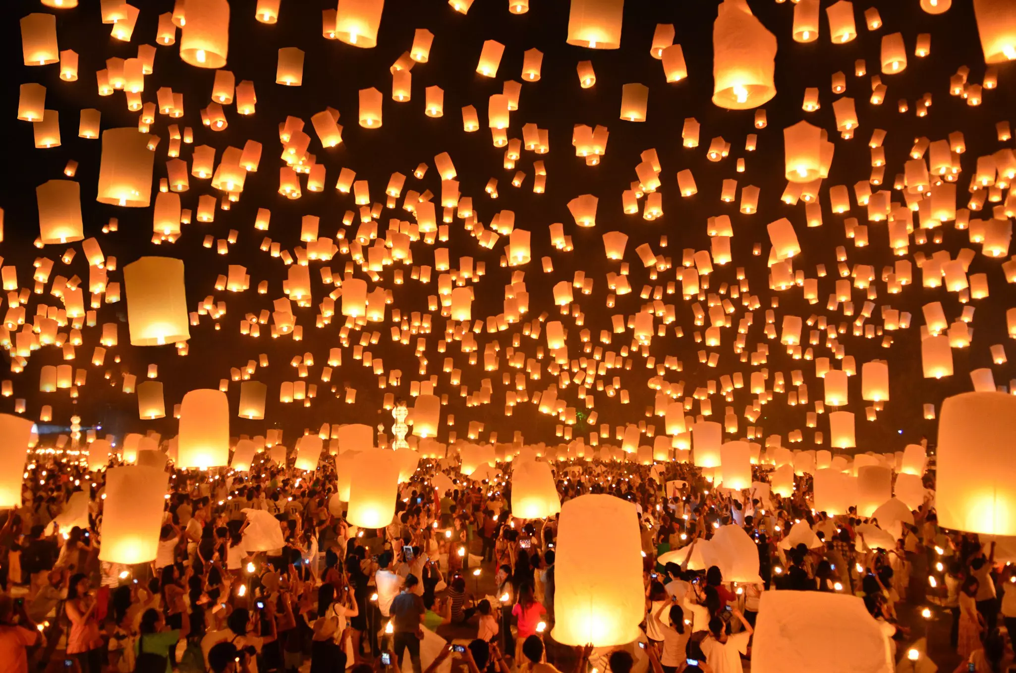 A huge crowd of people lighting paper lanterns and sending them up into the sky. Many people are holding cell phones and taking photographs